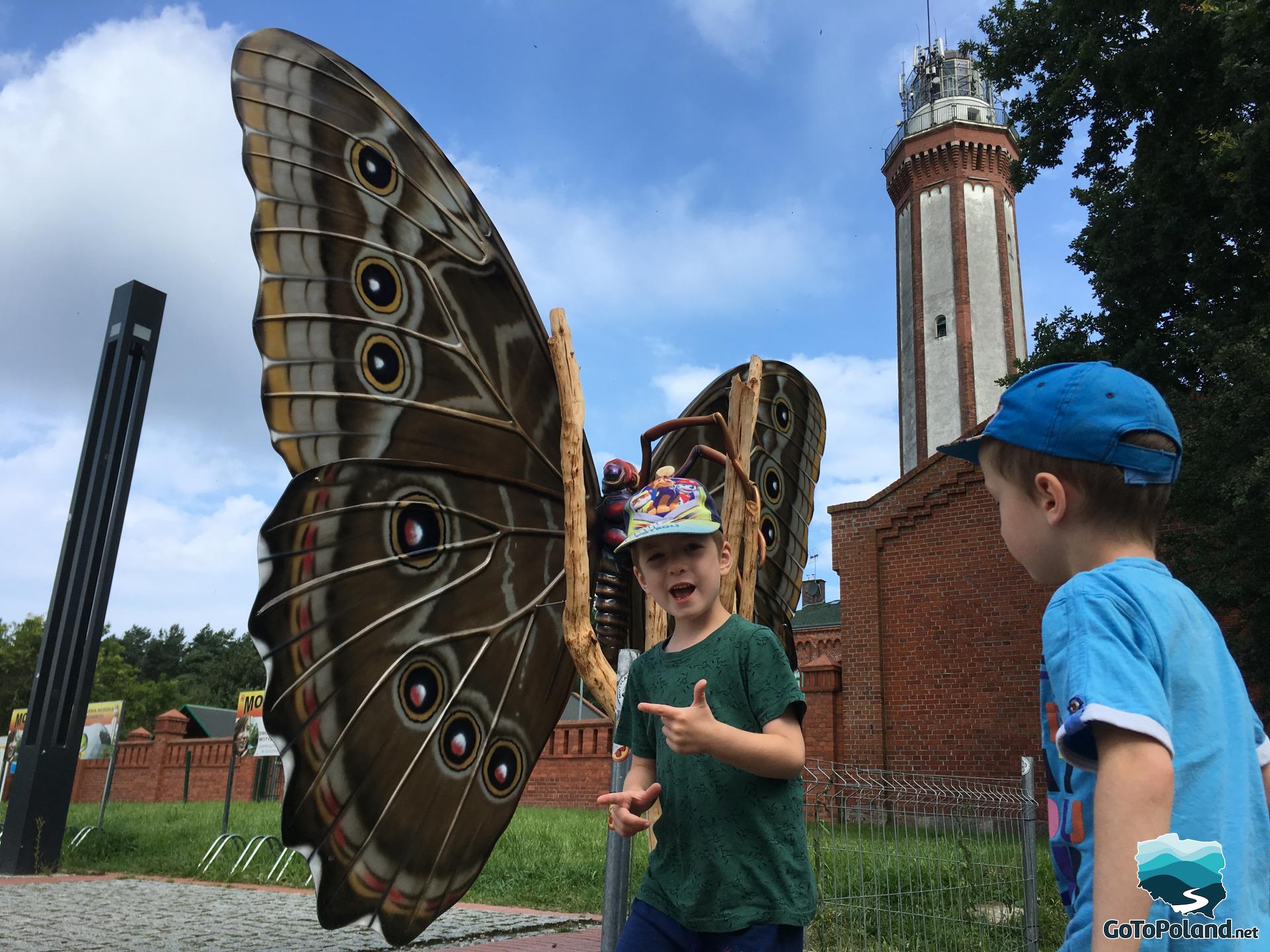 two boys in front of a big brown model of butterfly, behind them is a lighthouse