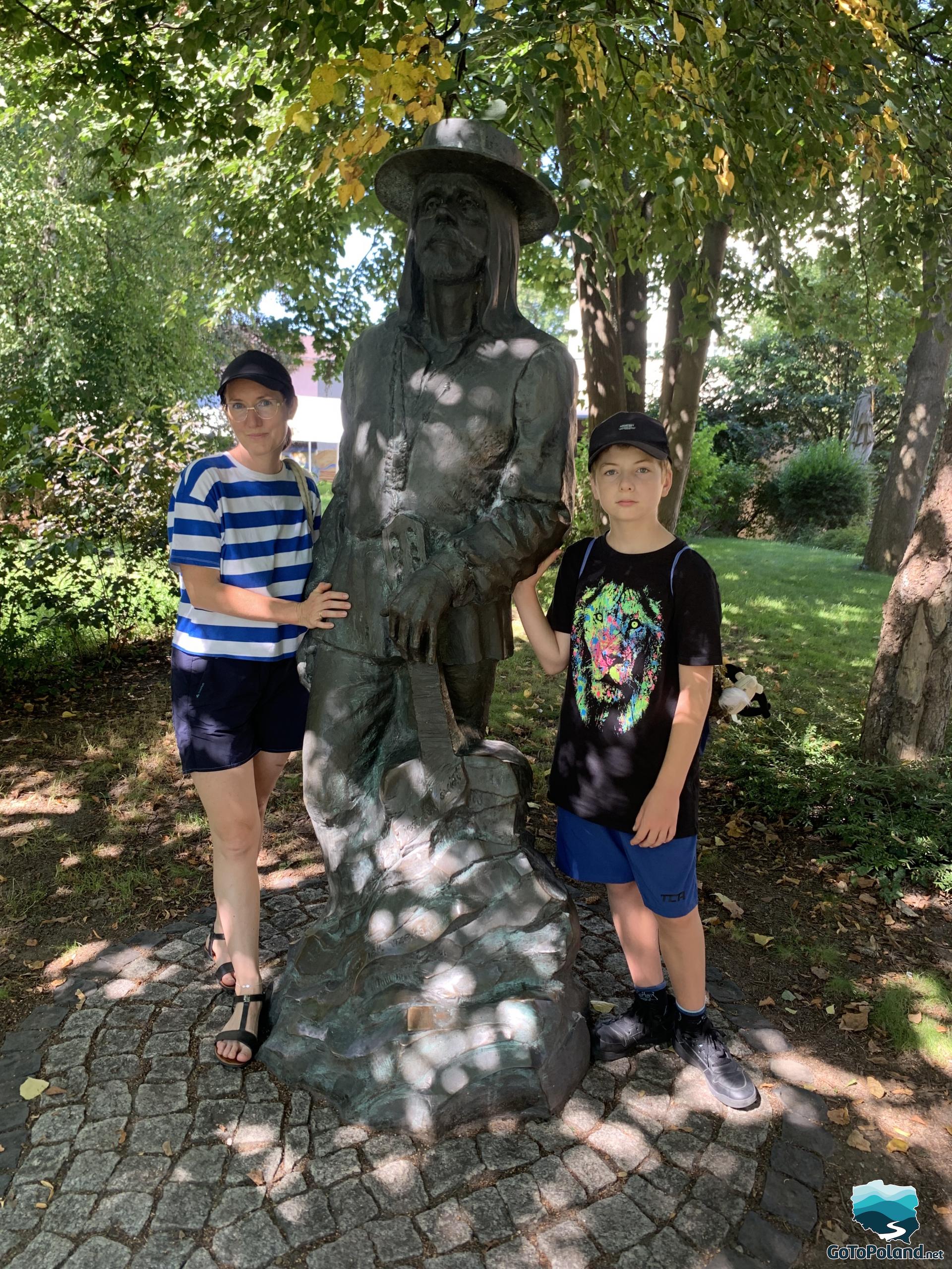 a sculpture of a famous Polish singer located in the shade of a tree, a woman and a boy are standing next to the sculpture