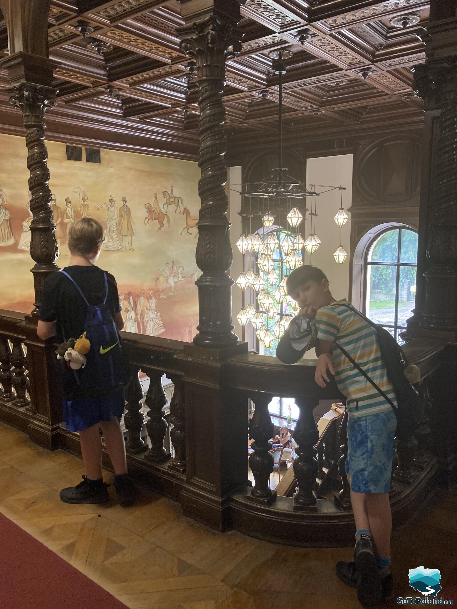 two boys leaning on the balustrades on the first floor of the castle, coffered ceiling