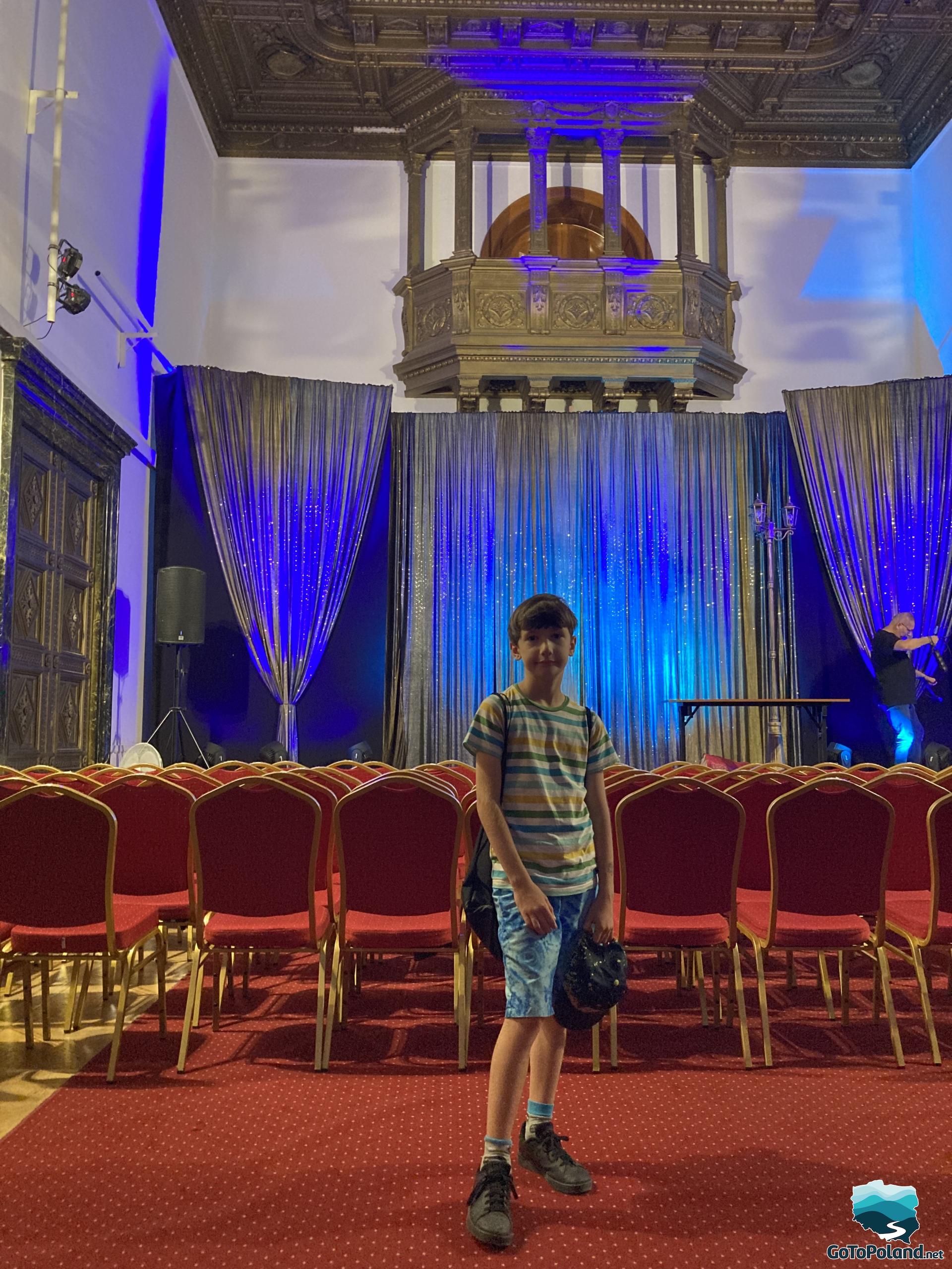the boy stands in the hall, behind him a blue curtain and rows of red chairs