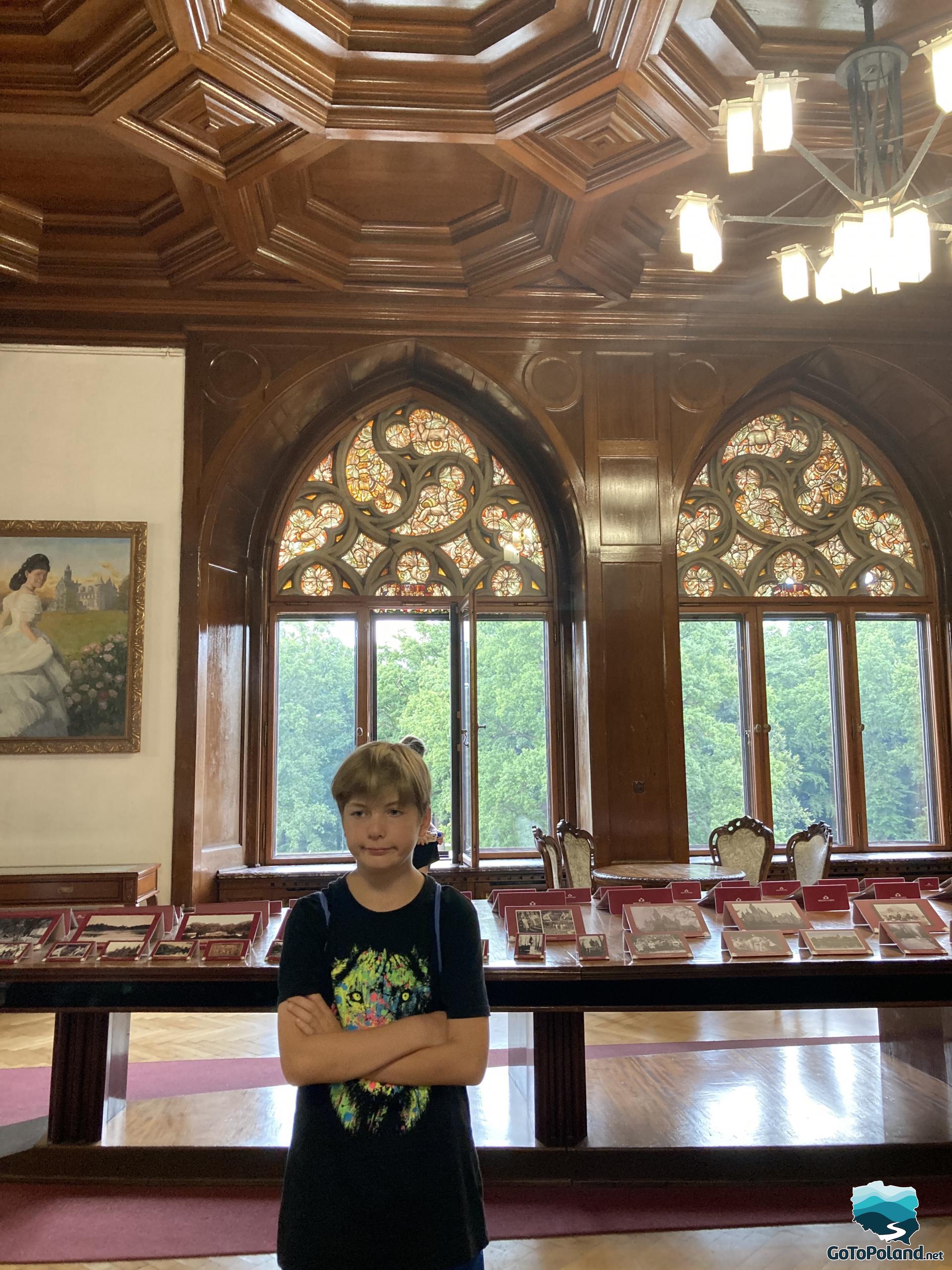 the boy stands in the hall, behind him is a table with postcards and two windows in the Gothic style