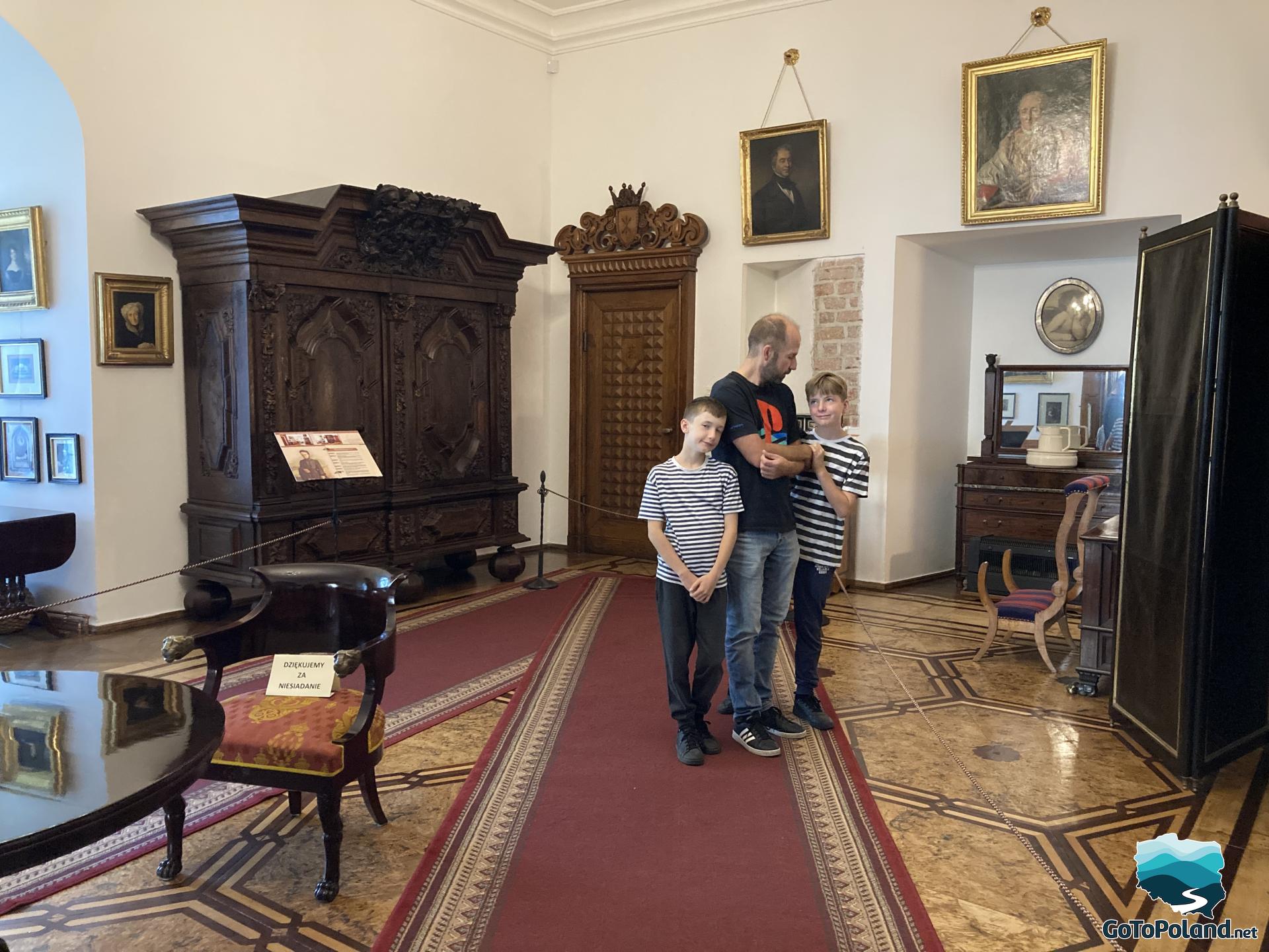 A man and two boys standing in a very ornate room with a wooden wardrobe, chairs and several paintings on the wall