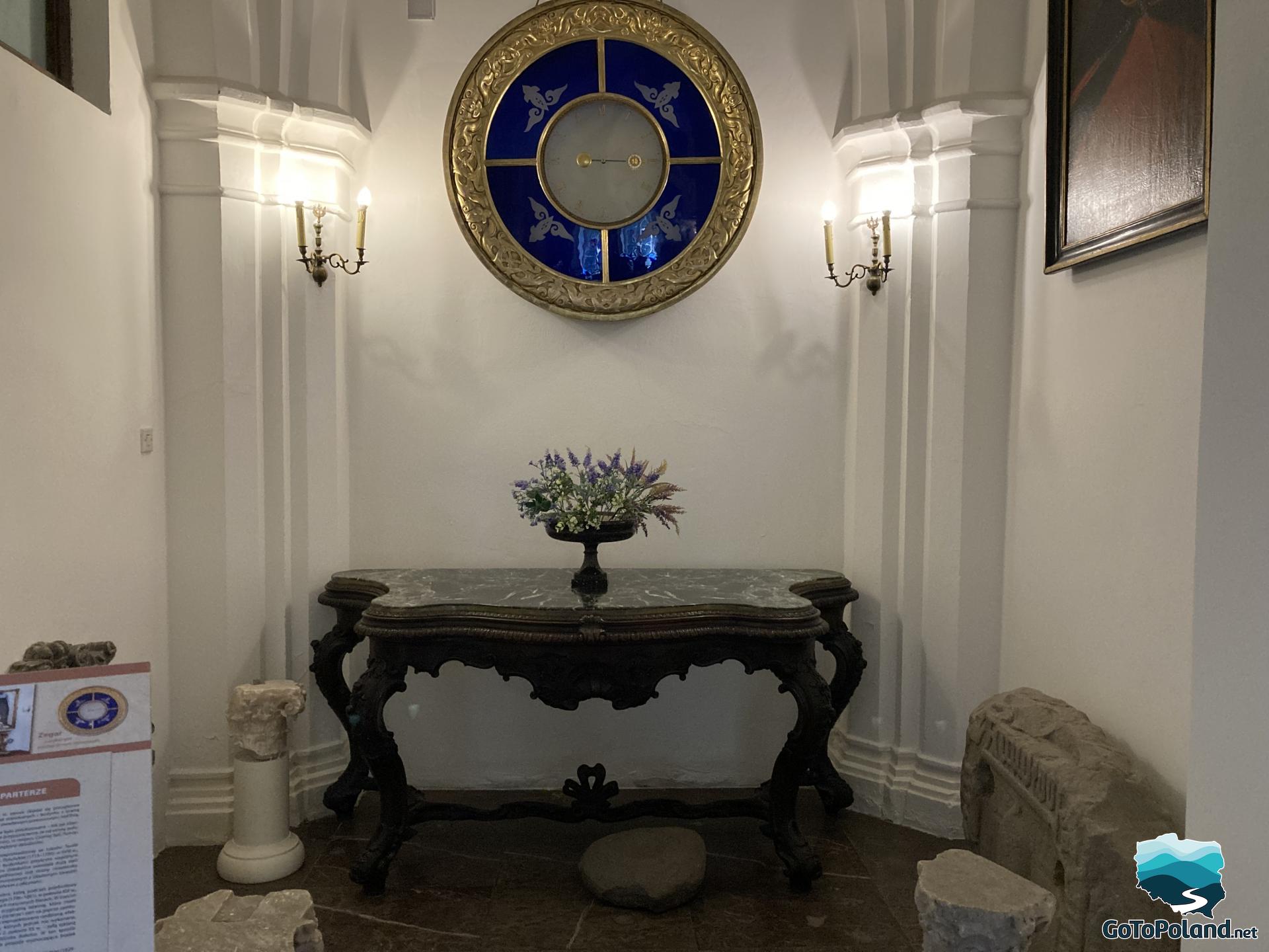 an antique table with flowers on it, a huge blue and gold clock on the wall