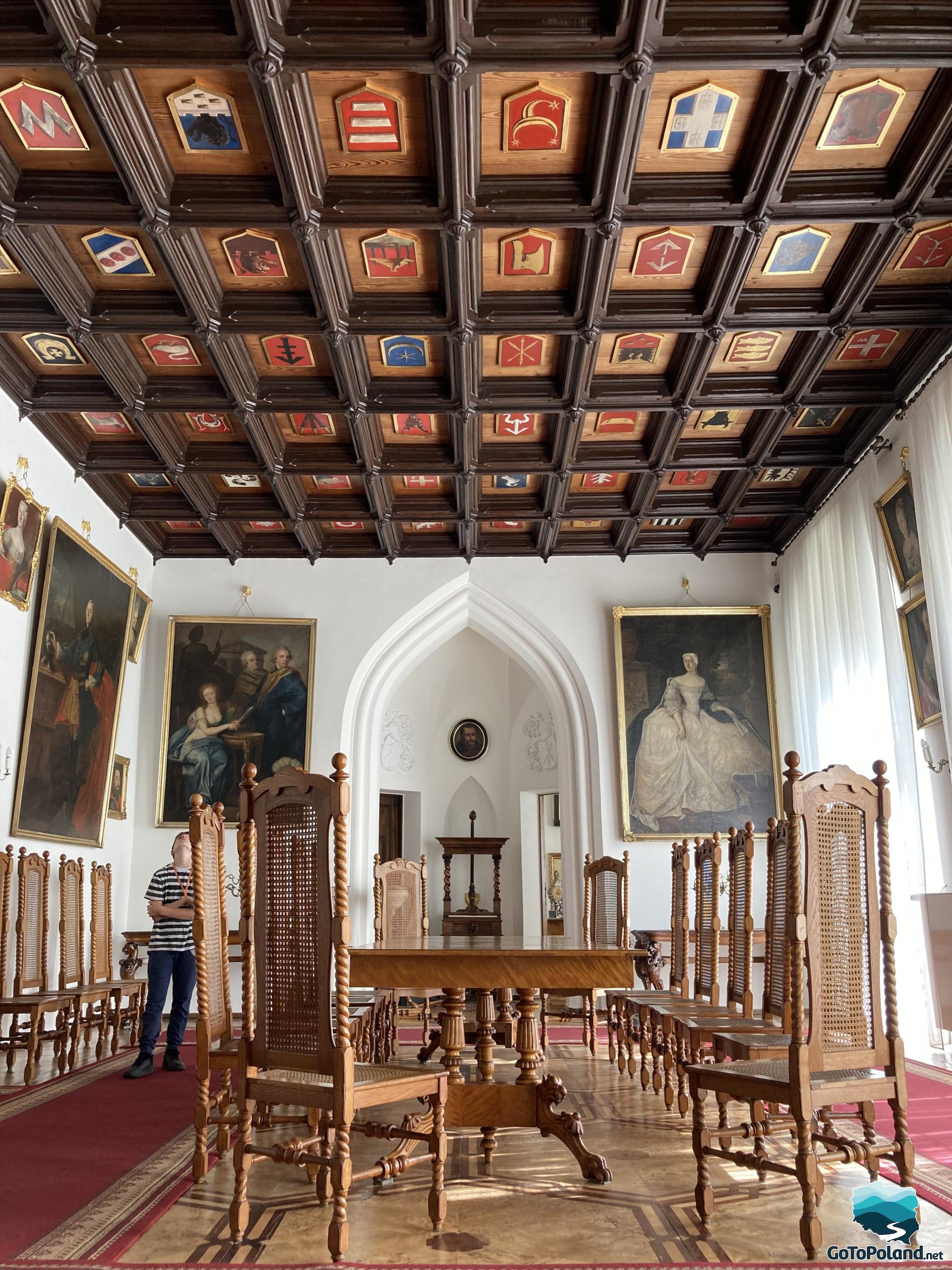a long wooden table, over ten chairs in a room, a boy is looking at the the coffered ceiling with coats of arms