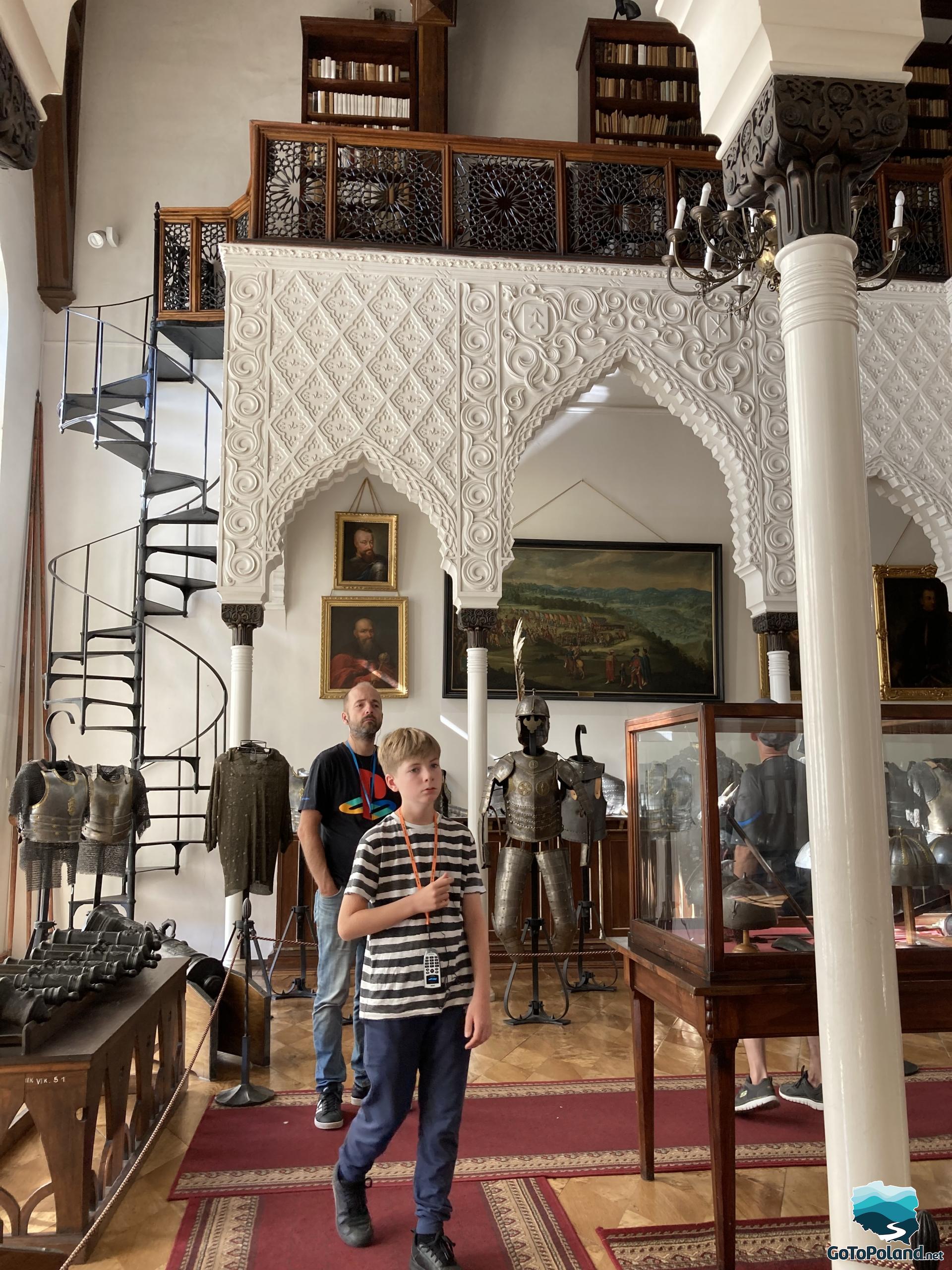 a man and a boy are looking at military souvenirs, gun barrels, armor, they are in a room whose architecture resembles Arabic patterns