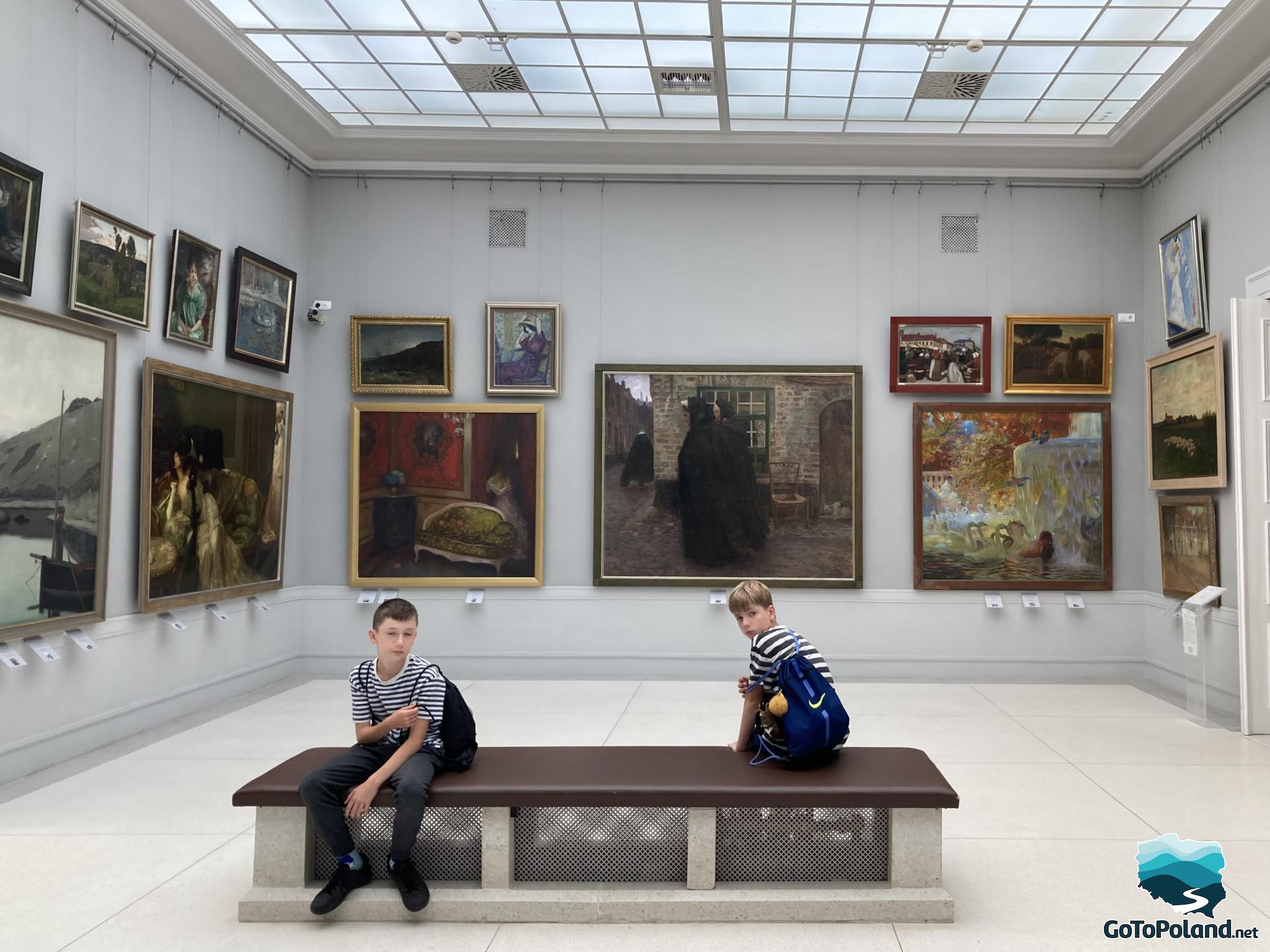 two boys are sitting in a painting gallery, there are lots of paintings hanging there