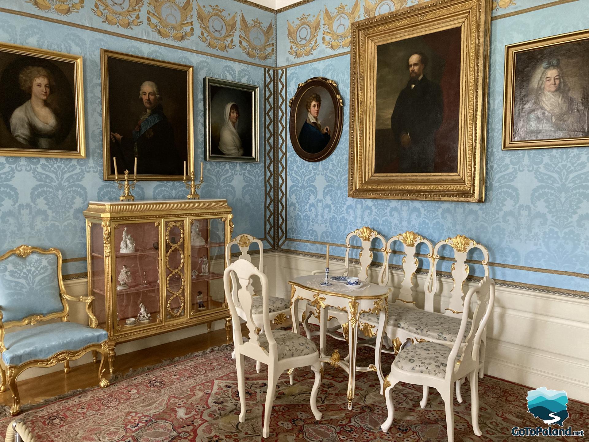 in the room there are white chairs, a blue armchair with gold legs, blue wallpaper and several portraits in gold frames on the walls