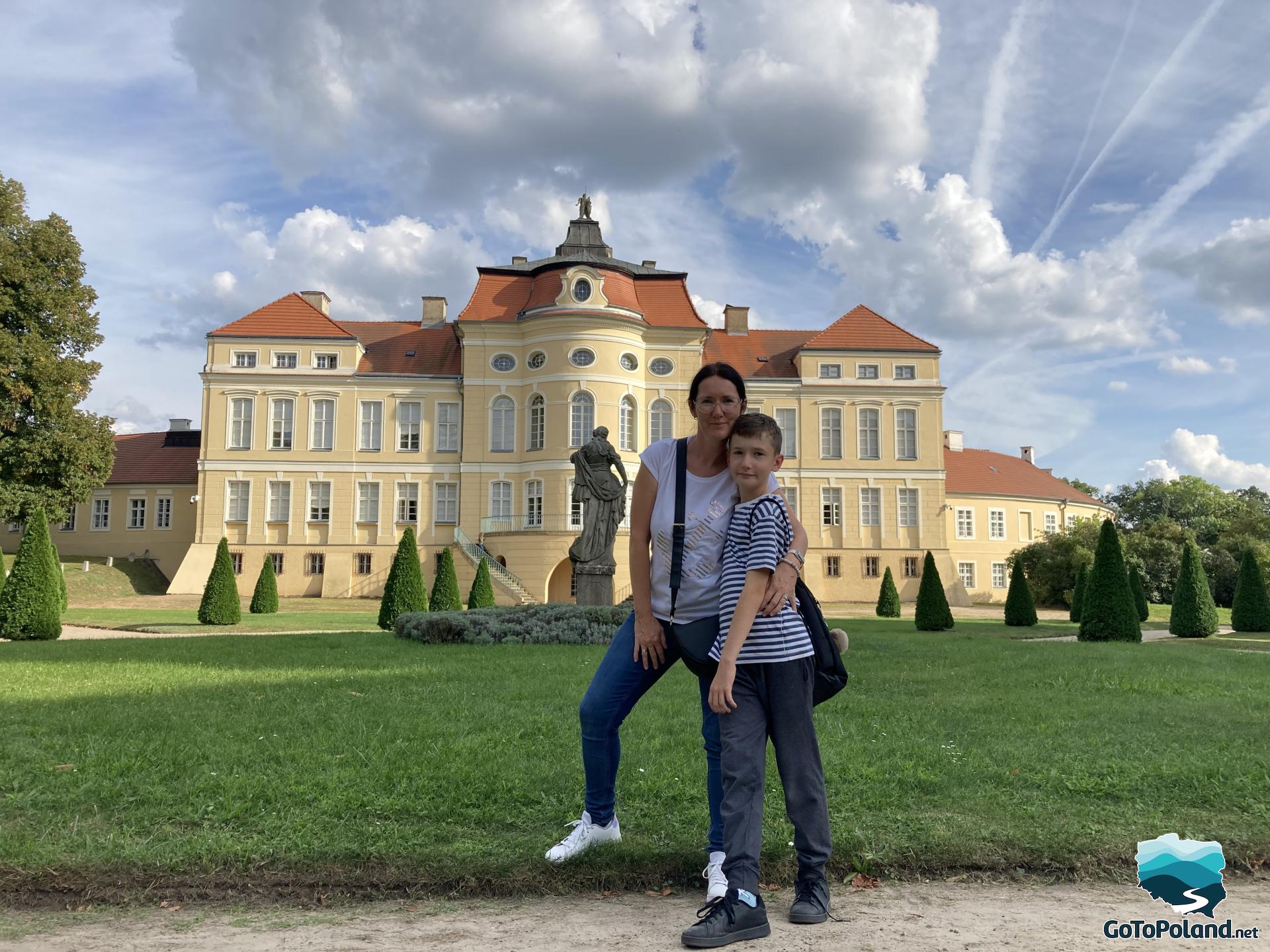 a woman and a boy are posing for a photo, behind them a lawn, a stone sculpture and a castle