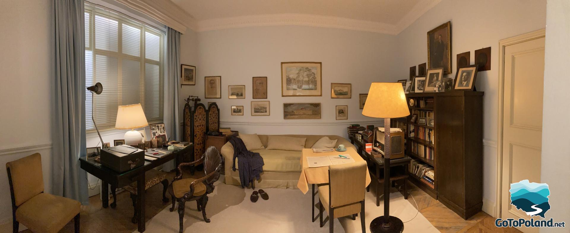 The room has a desk, a bed, two lamps, a shelf with books, a few photos, and a small table with chairs