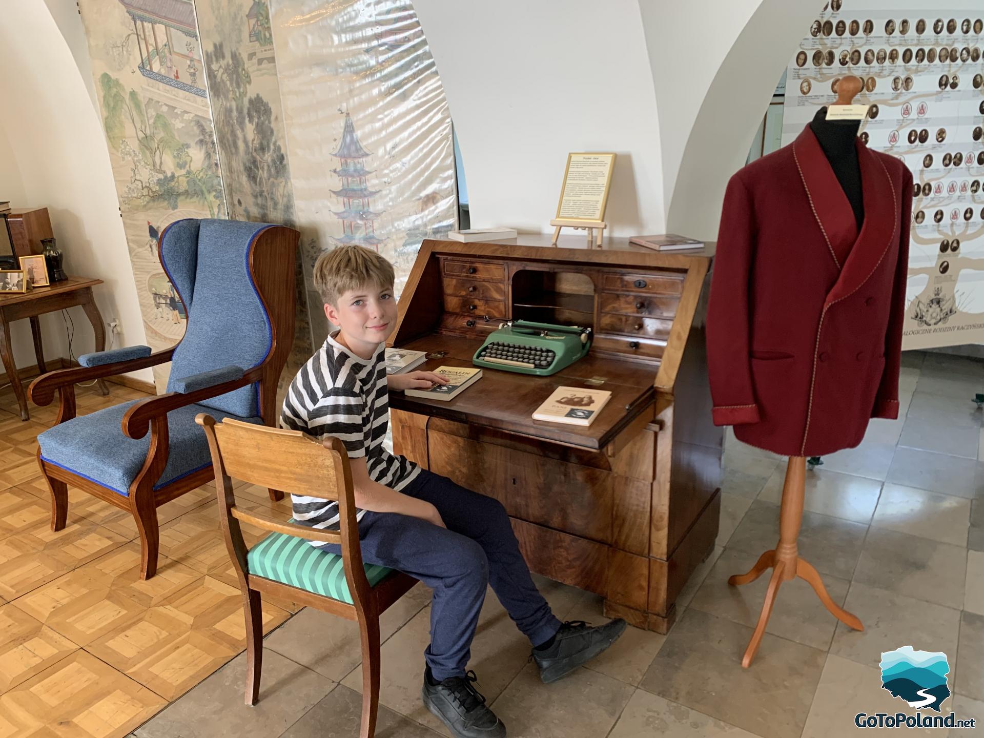 a boy is sitting next to a wooden desk, there is an old typewriter on the desk, next to it there is a hanger with a burgundy jacket