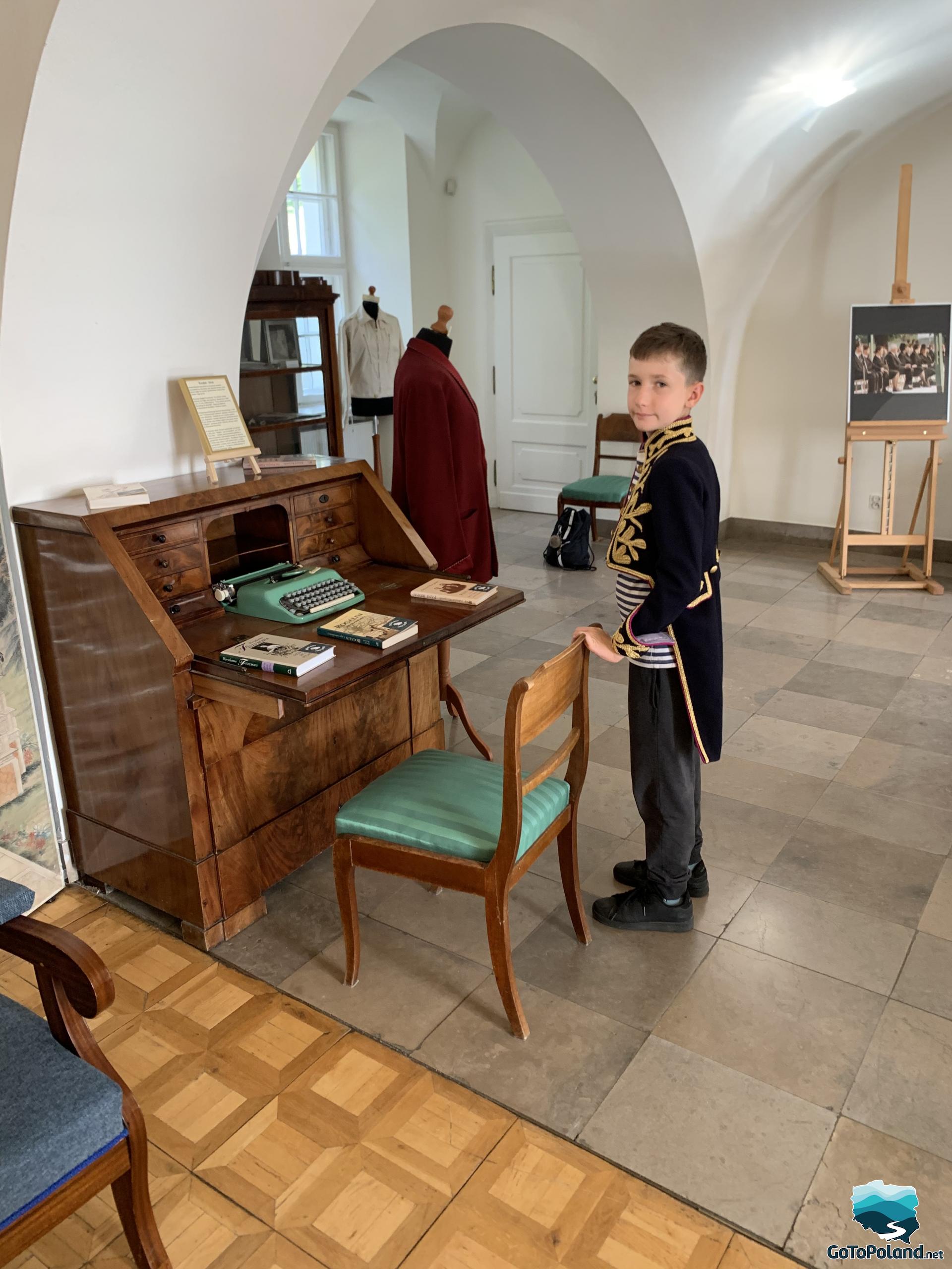 a boy is standing next to the wooden desk, he is wearing a tailcoat