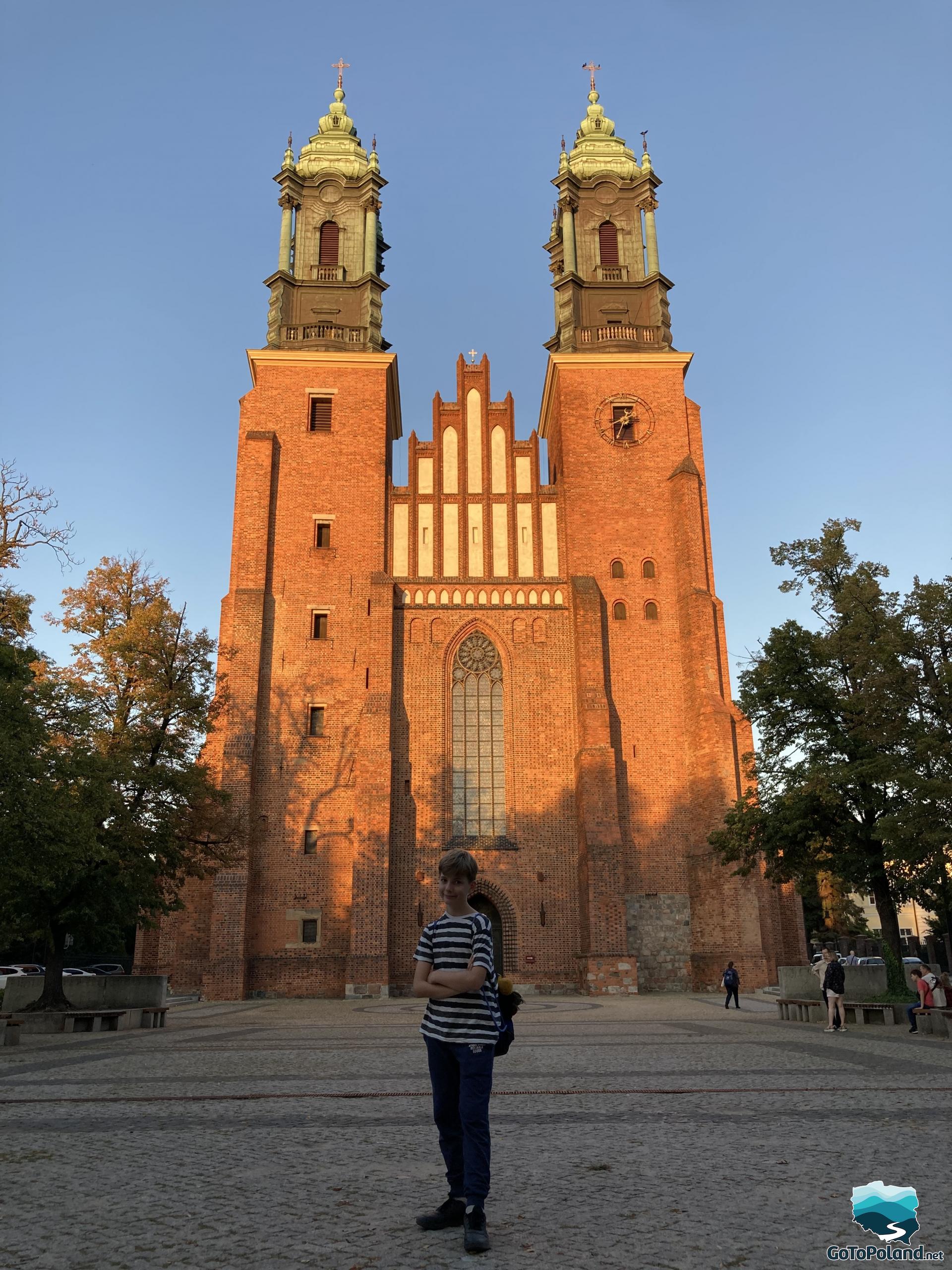 a boy standing in front of a tall red brick church with two huge towers