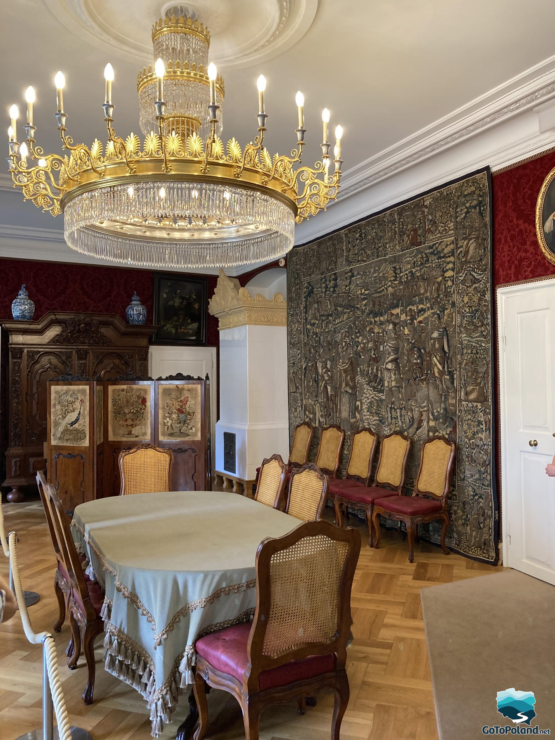 wooden chairs, a tapestry on the wall, a white stove, an oval table and a large gold crystal chandelier