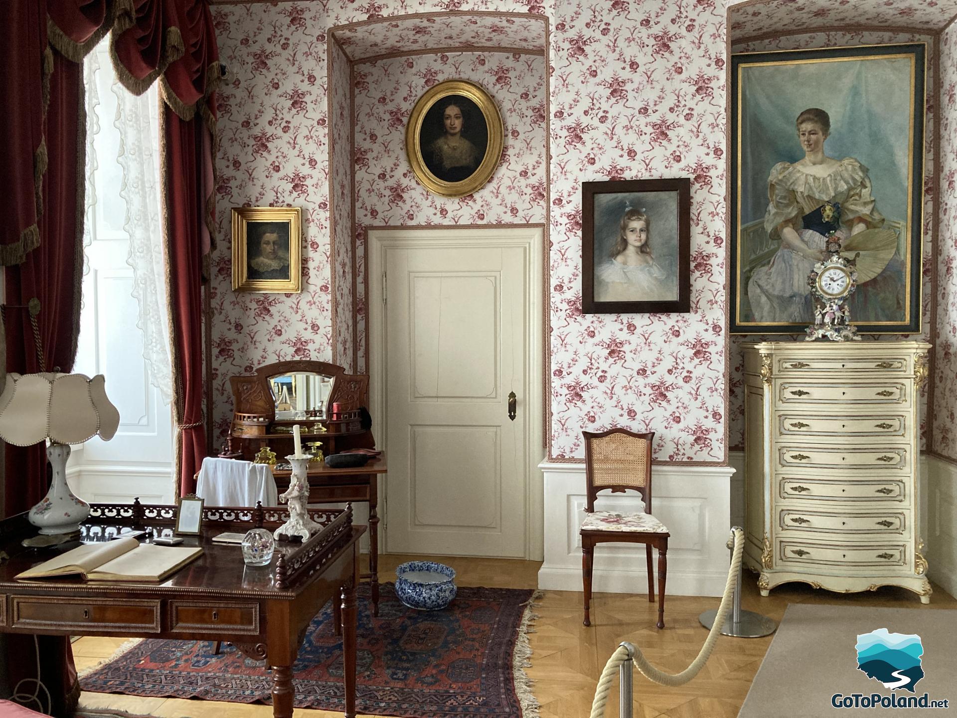 the room has a dressing table, a wooden desk, portraits on the walls and floral wallpaper on the walls