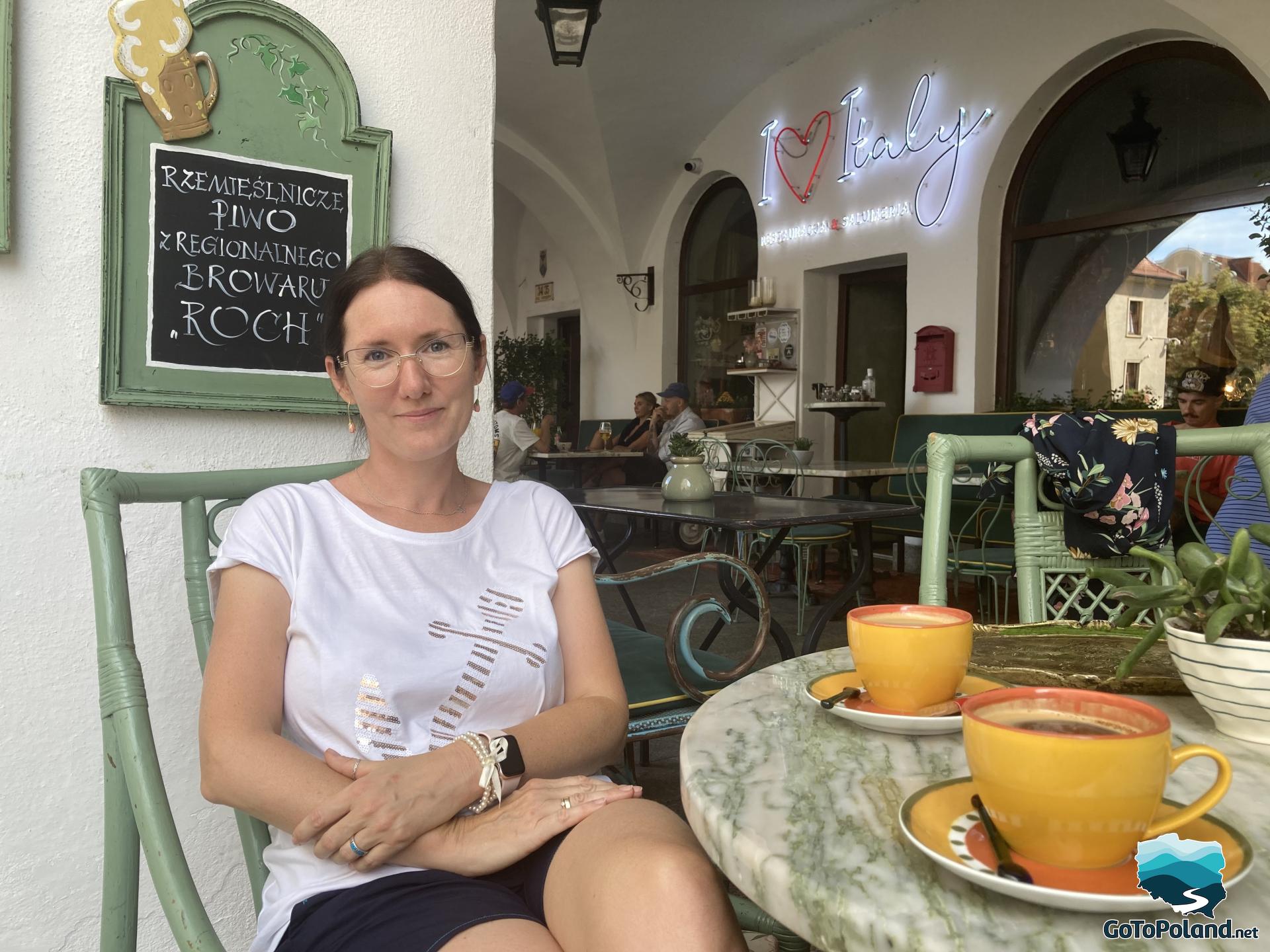 woman sits at a table in an outdoor cafe, two orange coffee cups stand on the table