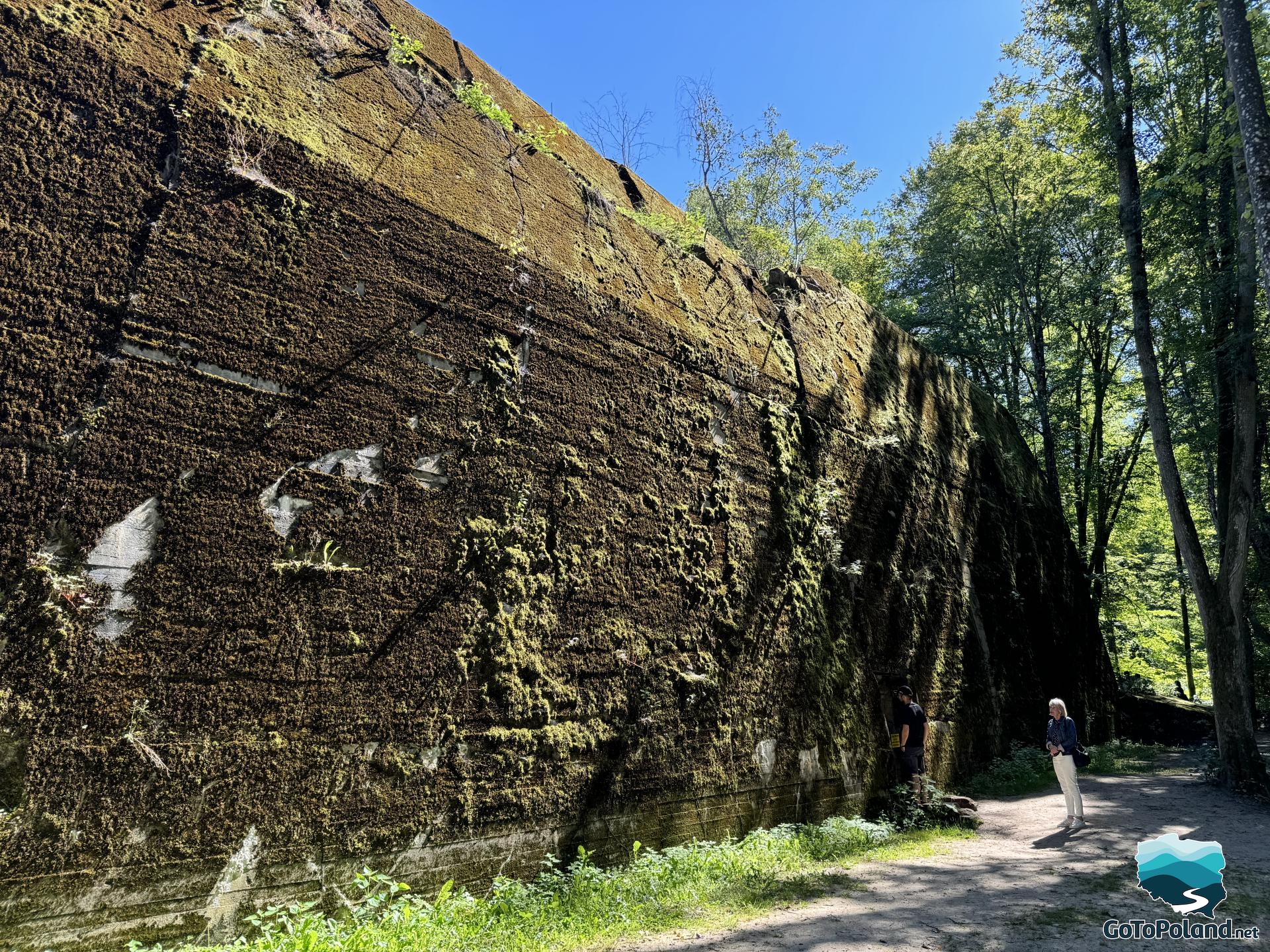 a woman stands in front of a huge wall (part of bunker)