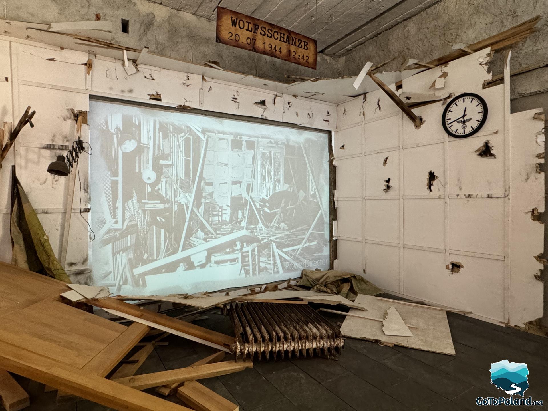 a room stylized as a room after a bomb explosion, with scattered boards on the floor, and a screen displaying archival photos on the wall