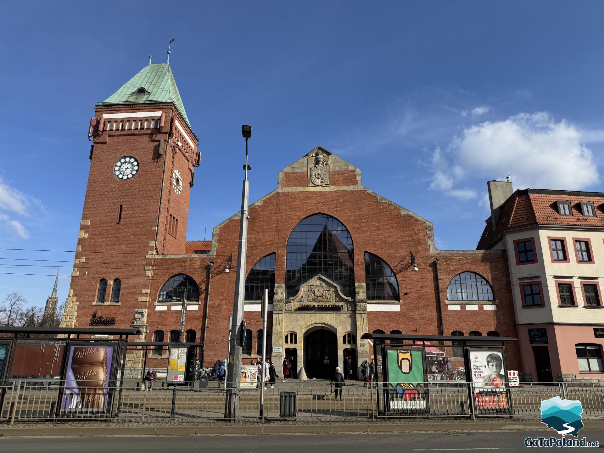 two tram stops and behind them a building with a brick tower, above the entrance to the building the date 1908