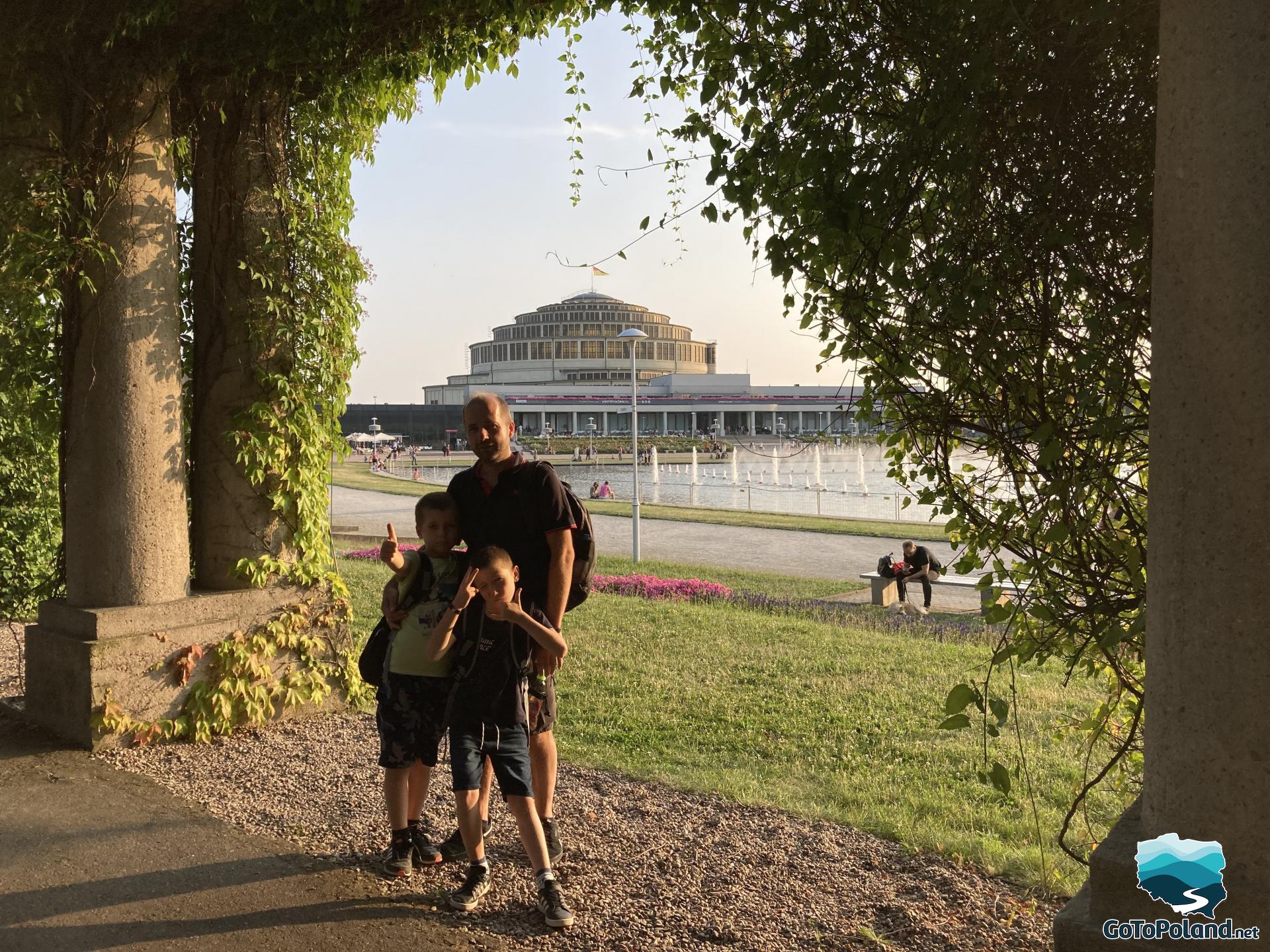 two children and a man are standing between a pergola, behind them a round fountain and a round building in the distance