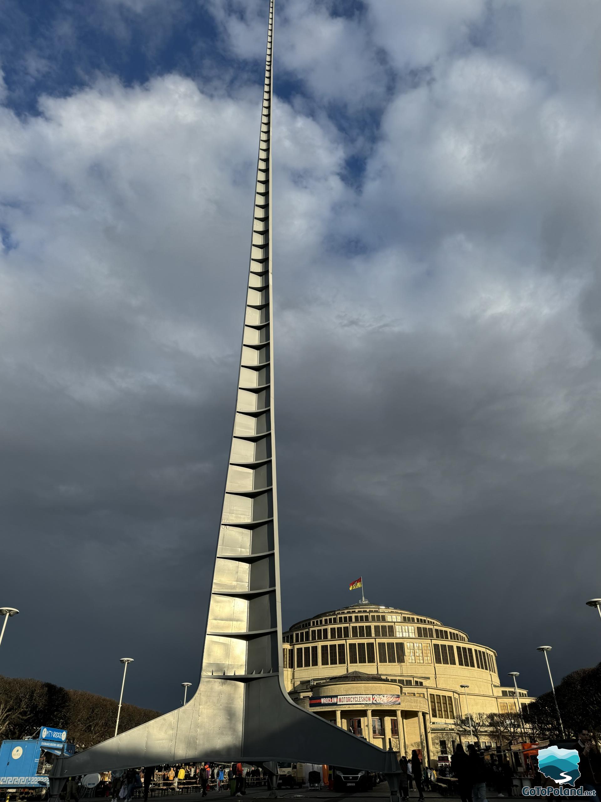 a tall steel spire, people around it, a round building behind it