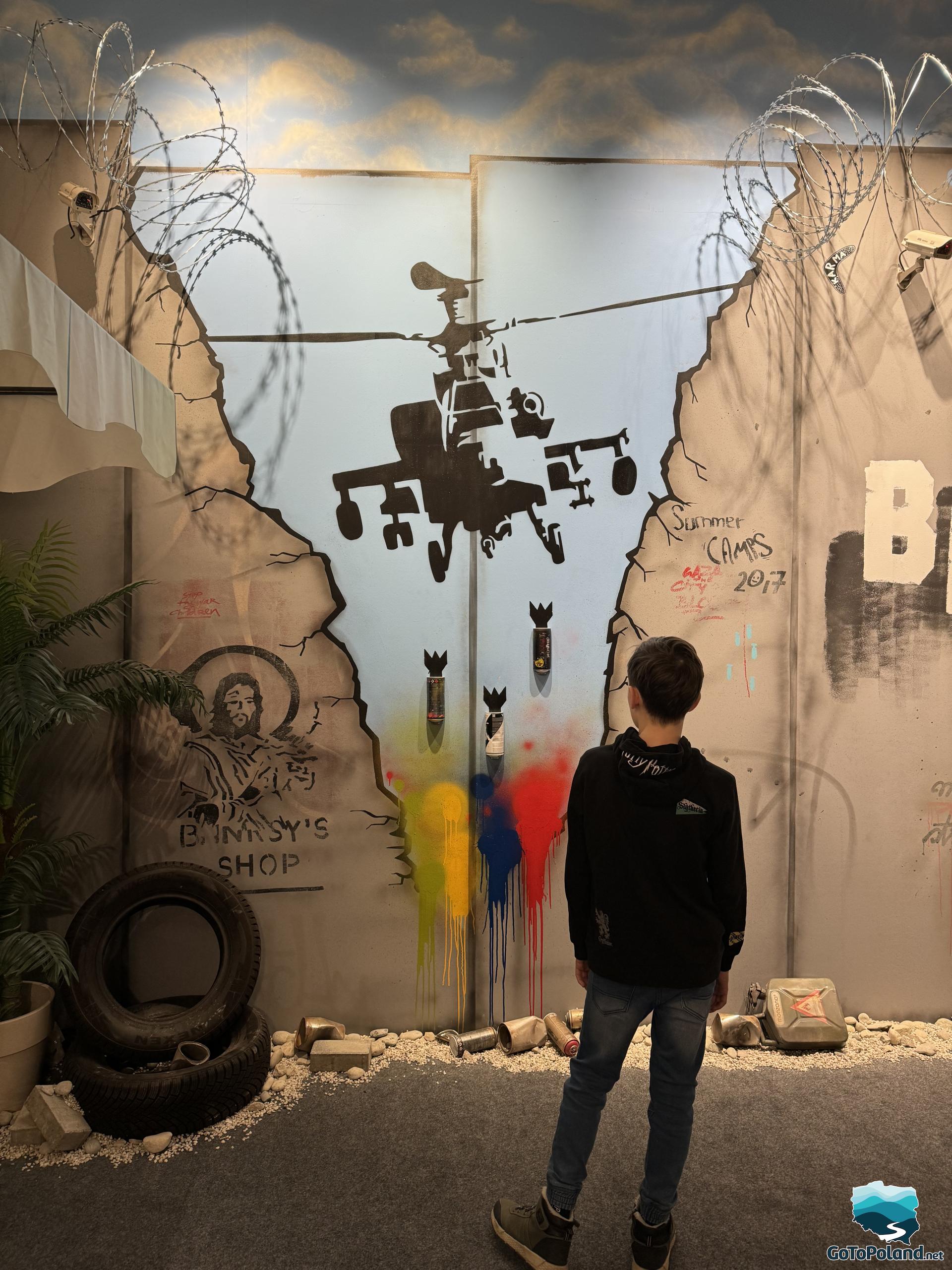 a boy looks at a work of art painted on the wall, its main elements are: a military helicopter dropping bombs, some crushed rubble by the wall and two black tires