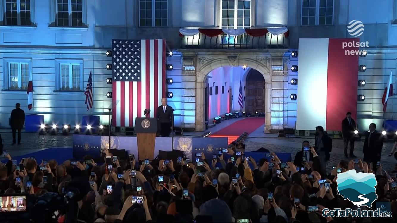 The President of the United States of America is heading to the rostrum in front of the castle, a crowd of people is gathered in front of the president, it is evening, flags hang on both sides of the entrance to the castle: one USA, the other Polish
