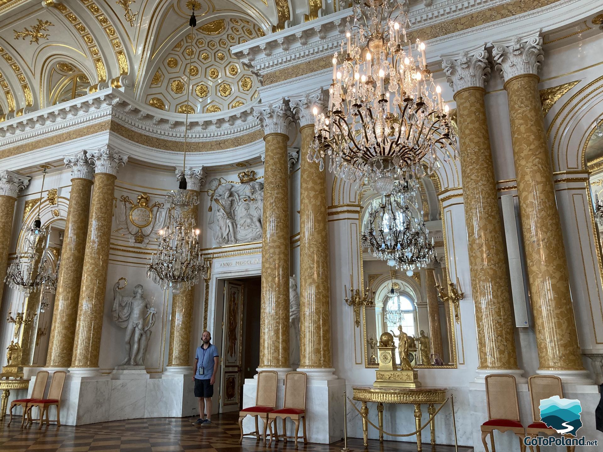 a ceremonial ballroom in the castle, a checkerboard floor, crystal chandeliers, yellow columns with Corinthian capitals, Roman style sculptures, big mirror, golden table under the mirror