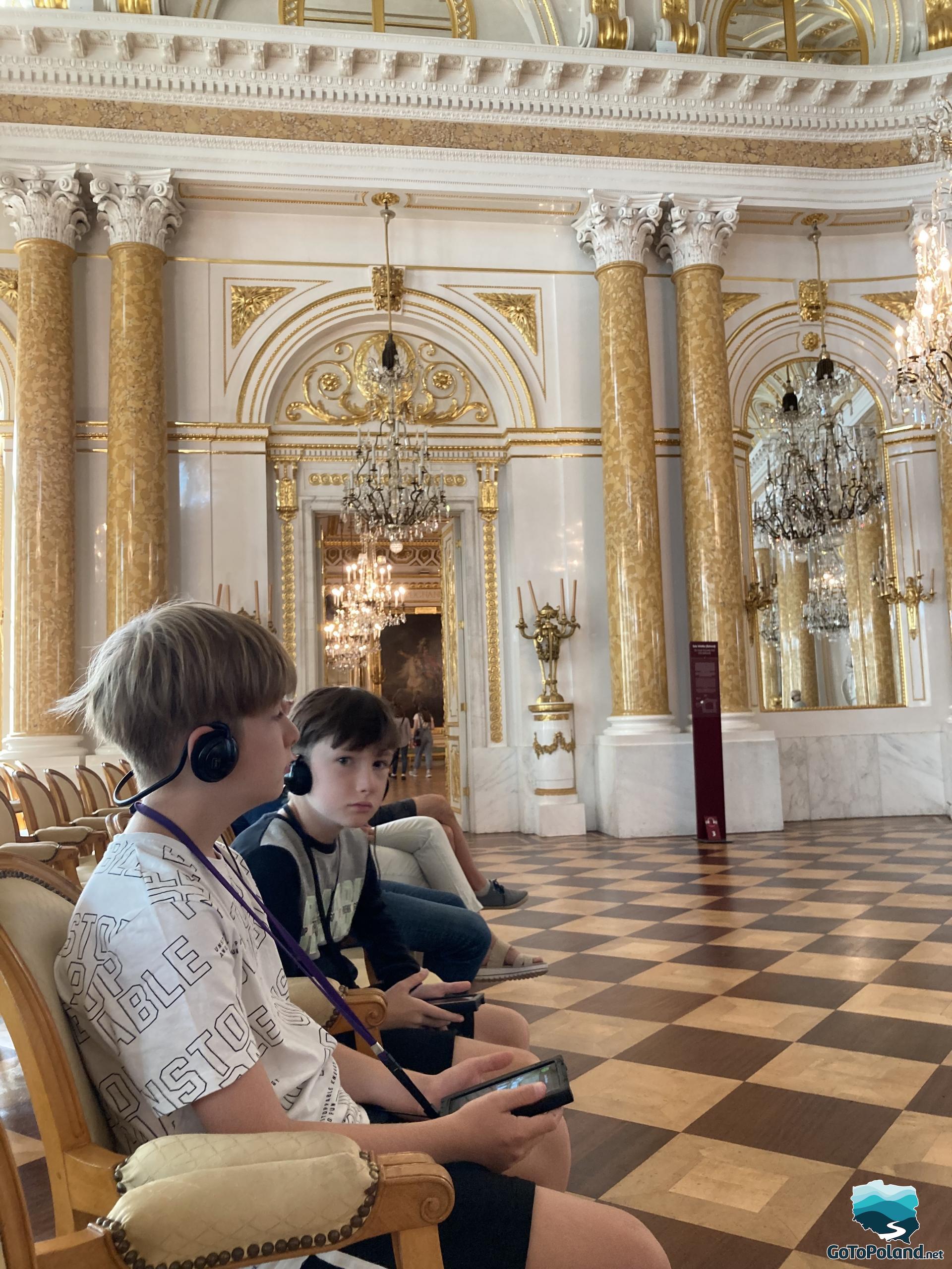two boys are sitting in the ballroom, they have audioguides in their hands and headphones on their ears, the hall is decorated with Roman-style columns and huge crystal chandeliers