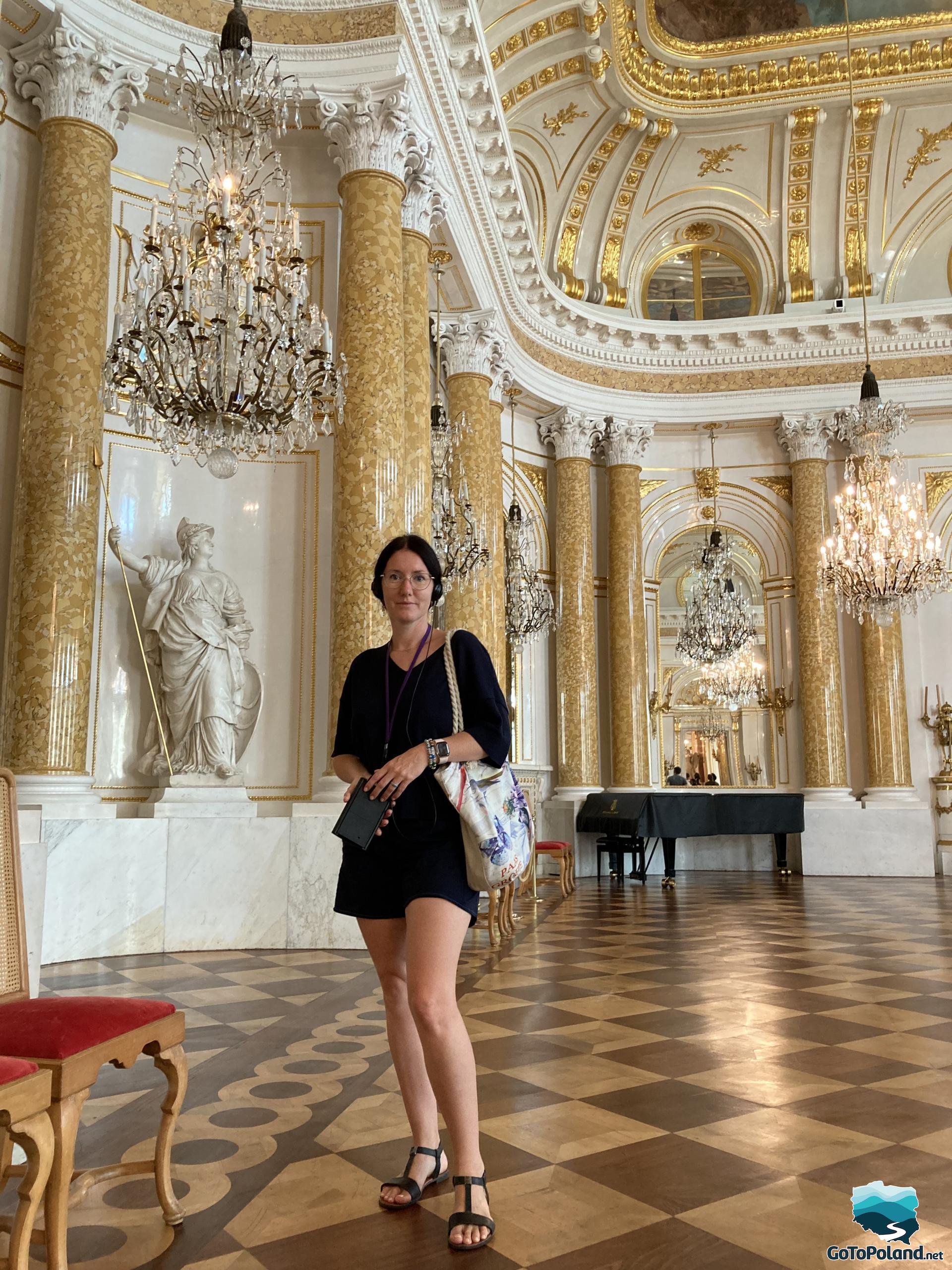 a woman is standing in the ballroom, behind her a black piano, you can see a fragment of a painted ceiling, the decoration of the room is Roman columns and crystal chandeliers and a sculpture