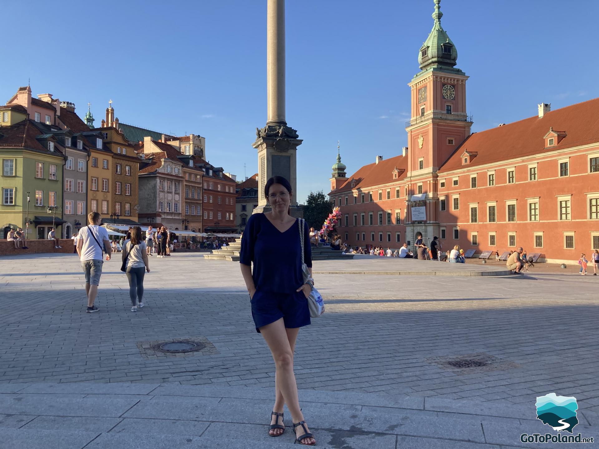 a woman is standing in the square, on the left there are colorful tenement houses, behind the woman there is a column, on the right there is a brick-red castle