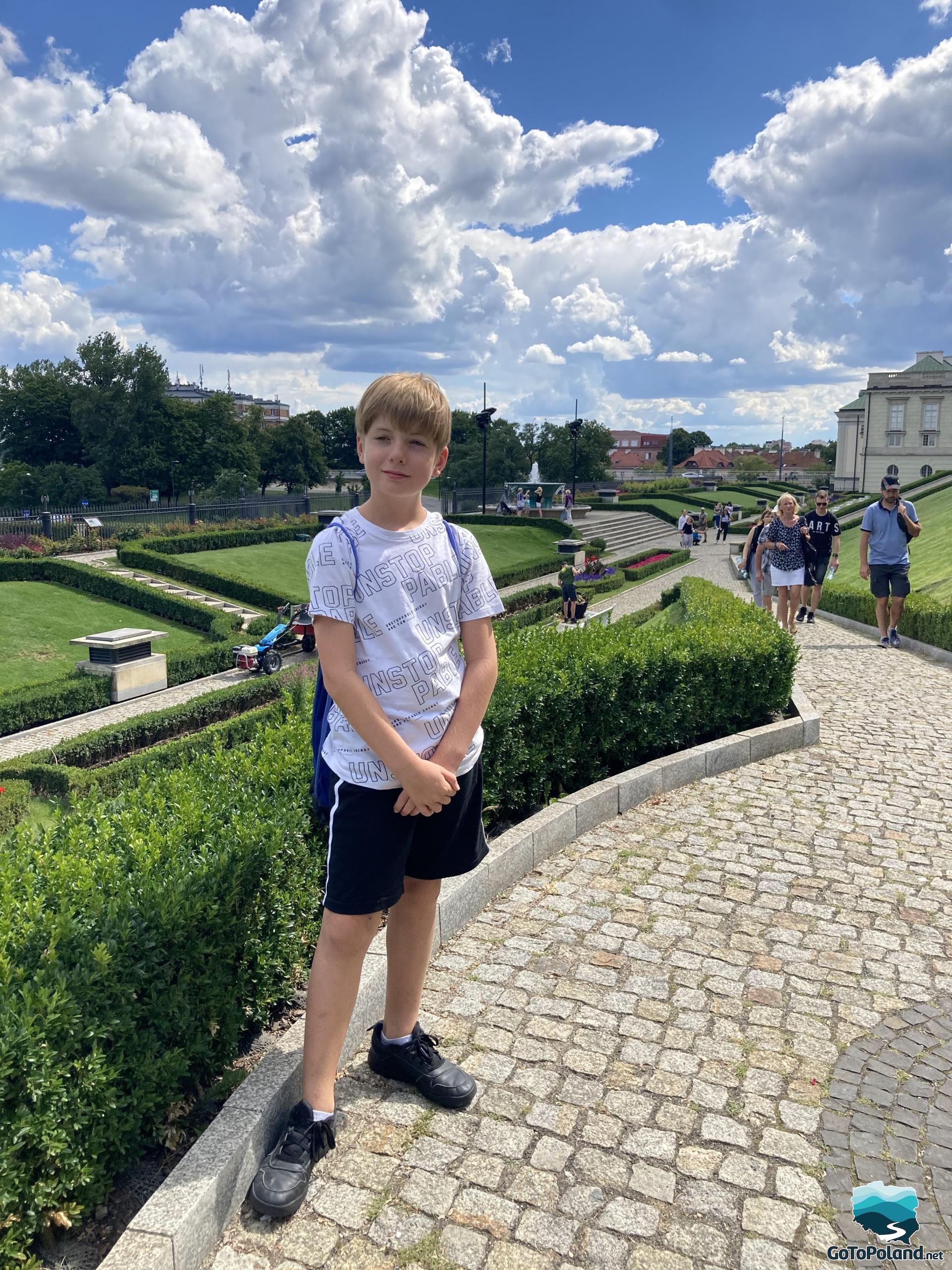 a boy in a white t-shirt and black shorts stands on a path made of cobblestones, the royal gardens stretch behind him