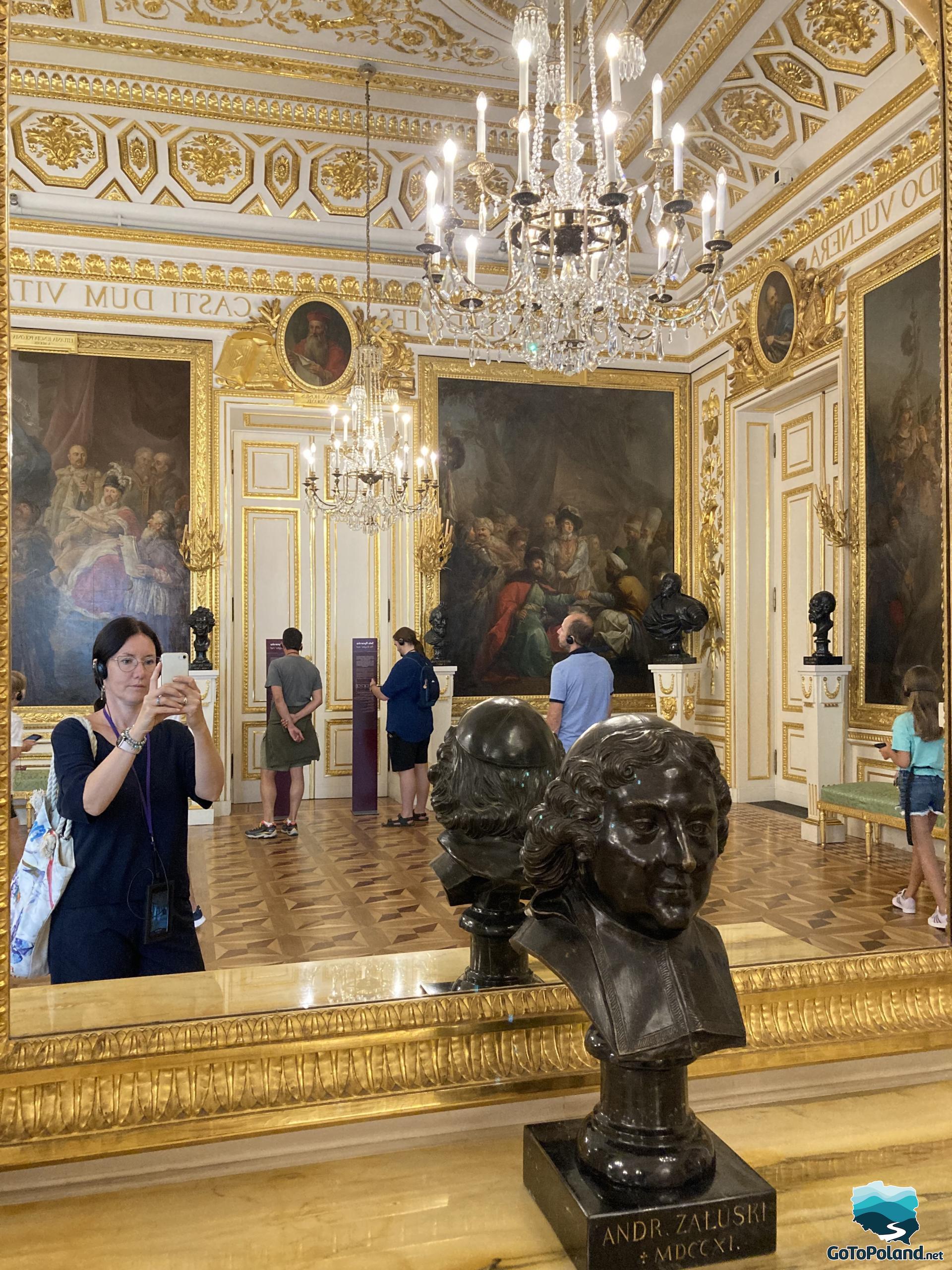 a woman is standing in front of a mirror and taking a picture looking at herself, the interior of the room with huge paintings, golden ornaments and crystal chandeliers is reflected in the mirror, a small black bust of a man stands in front of the mirror