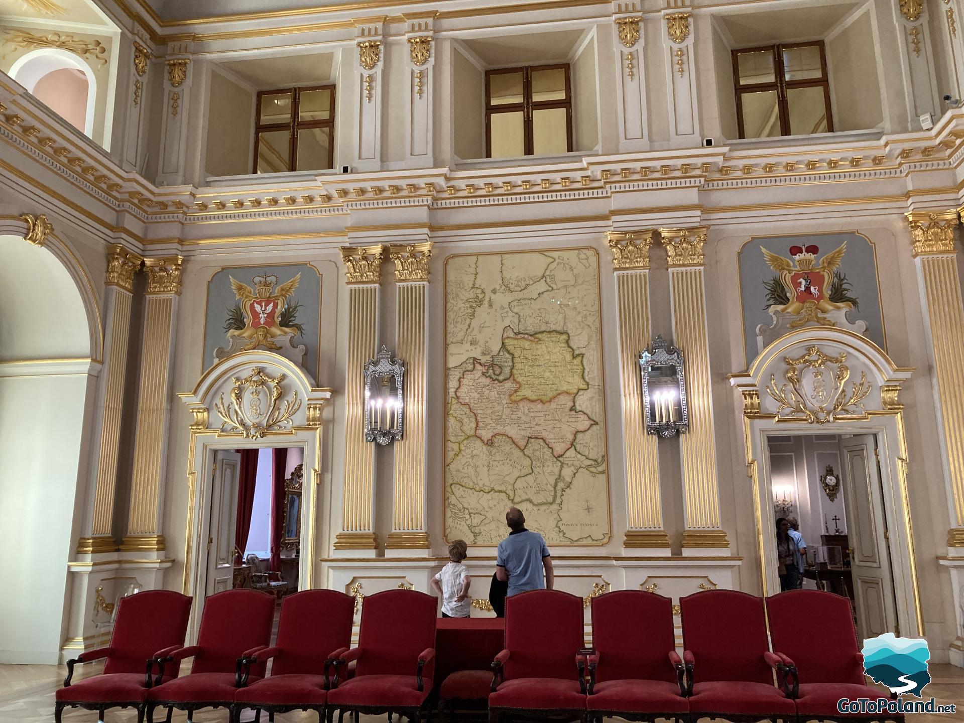 a man and two boys are looking at a map of former Poland with marked borders, on both sides of the map there are entrances to the hall, red chairs are in front,
