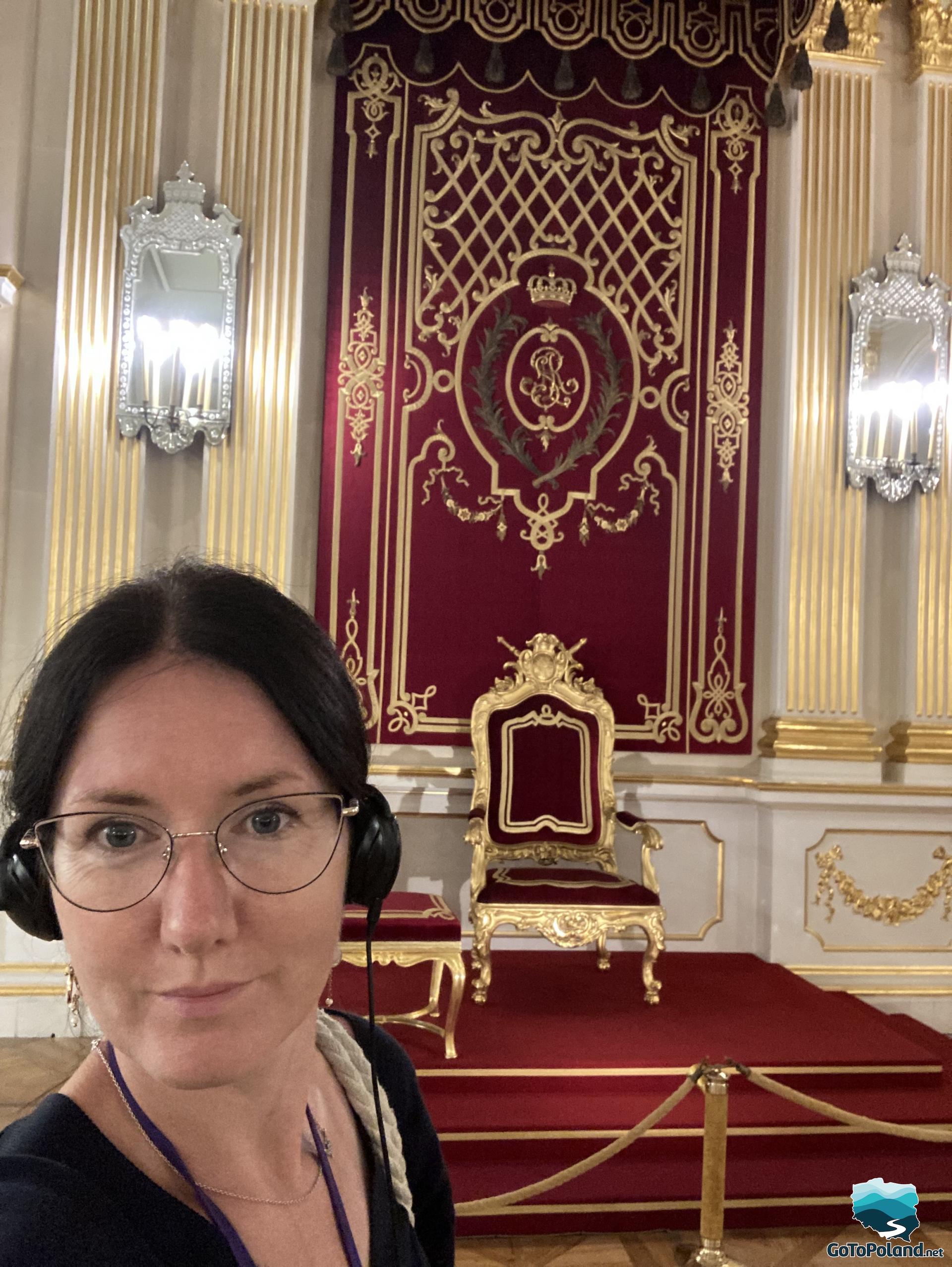 a woman is taking a selfie, behind her is a red throne chair with a gold frame, behind the throne chair is a wall with red fabric and a royal monogram on it