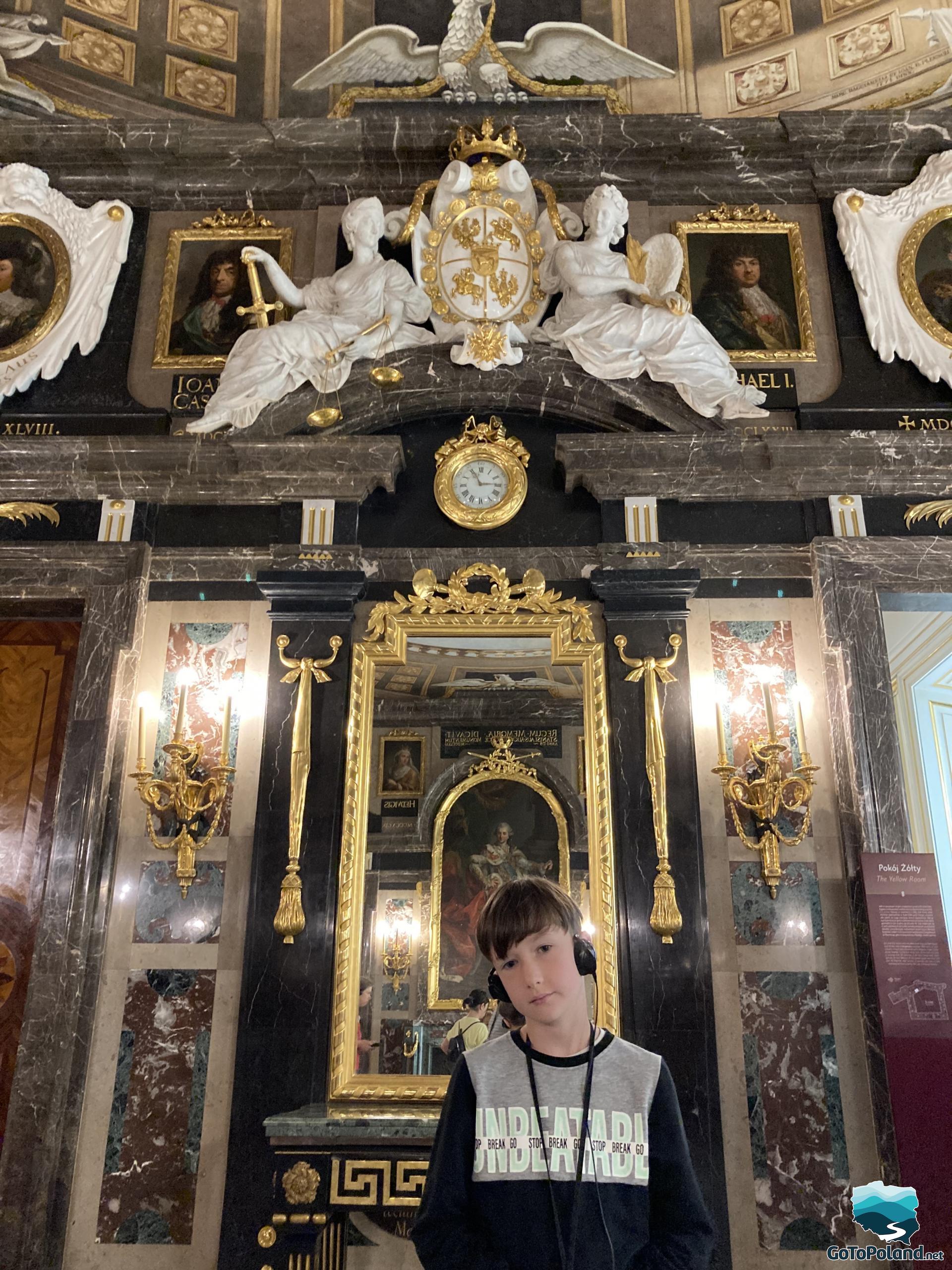 the boy is standing in a richly decorated room, there is a mirror behind it, white sculptures above the mirror, the walls in the room are made of marble