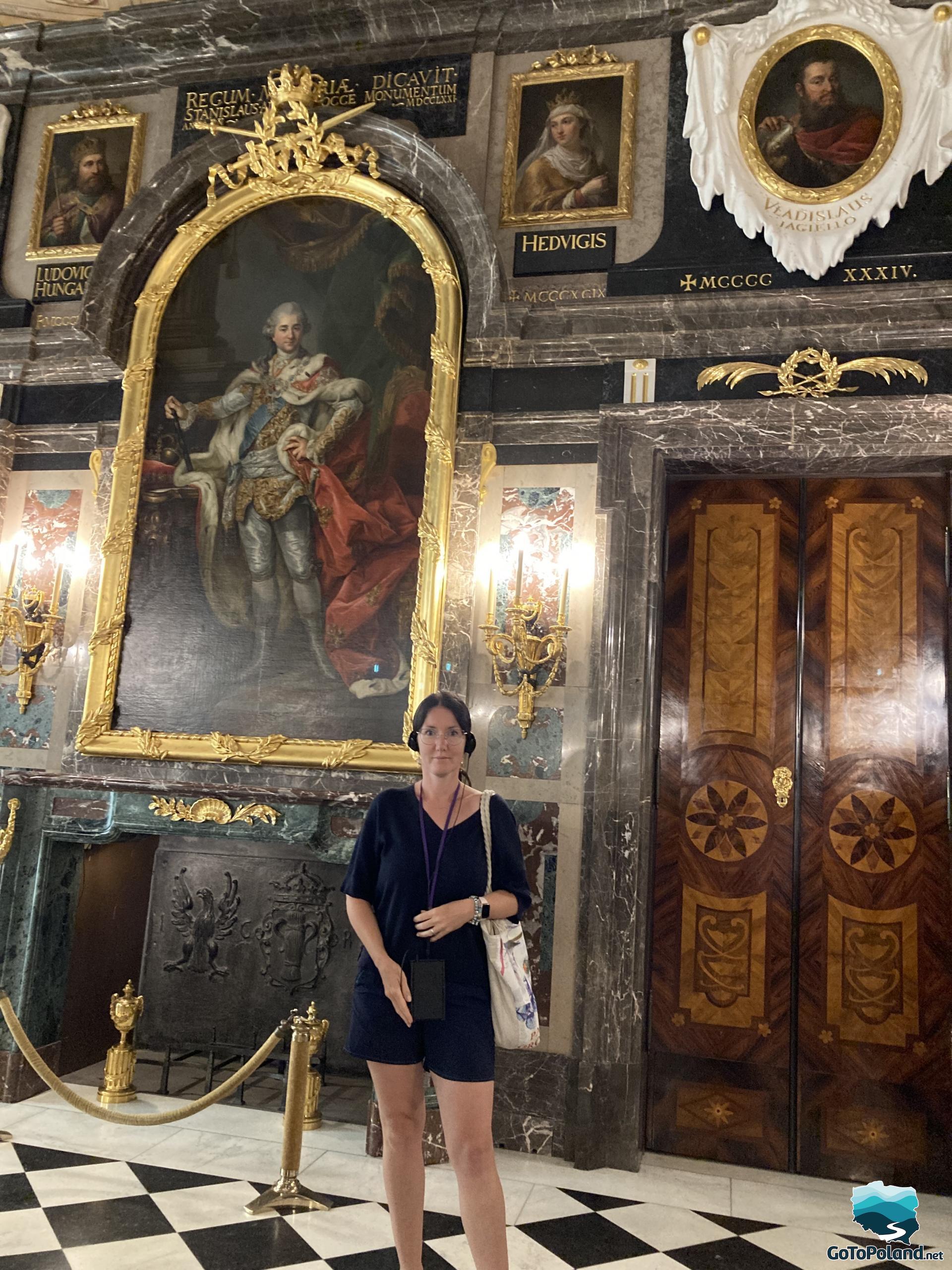 a woman is standing in a room with marble walls, a portrait of the last Polish king hangs behind her, there are images of other Polish rulers on the walls, you can also see inlaid doors