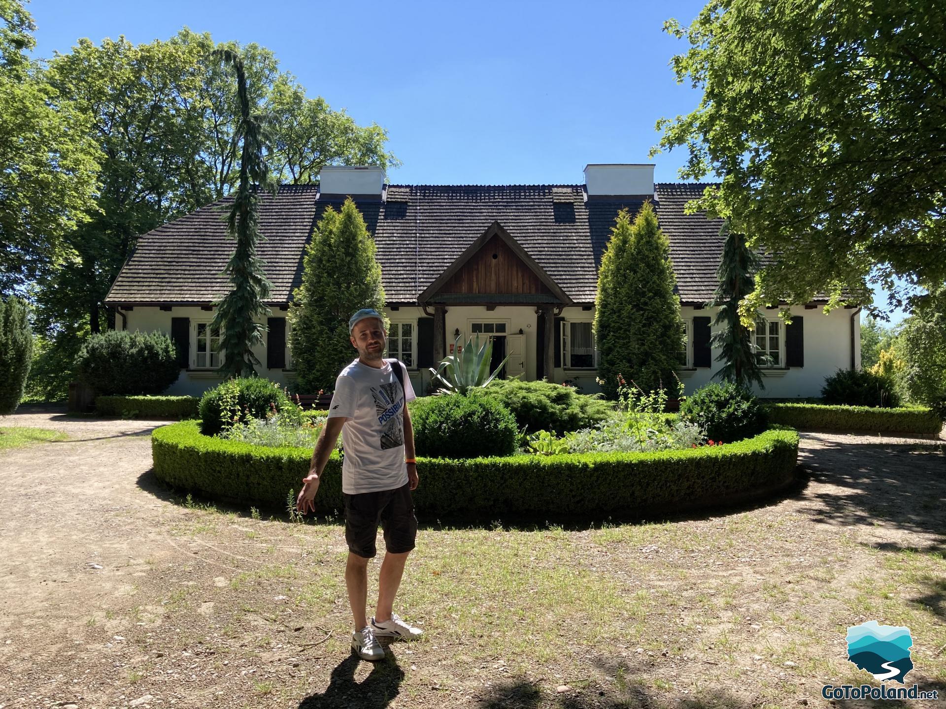 the man is standing in front of the manor house, there is a garden in the shape of a circle between the man and the manor house, and a circular path around the garden