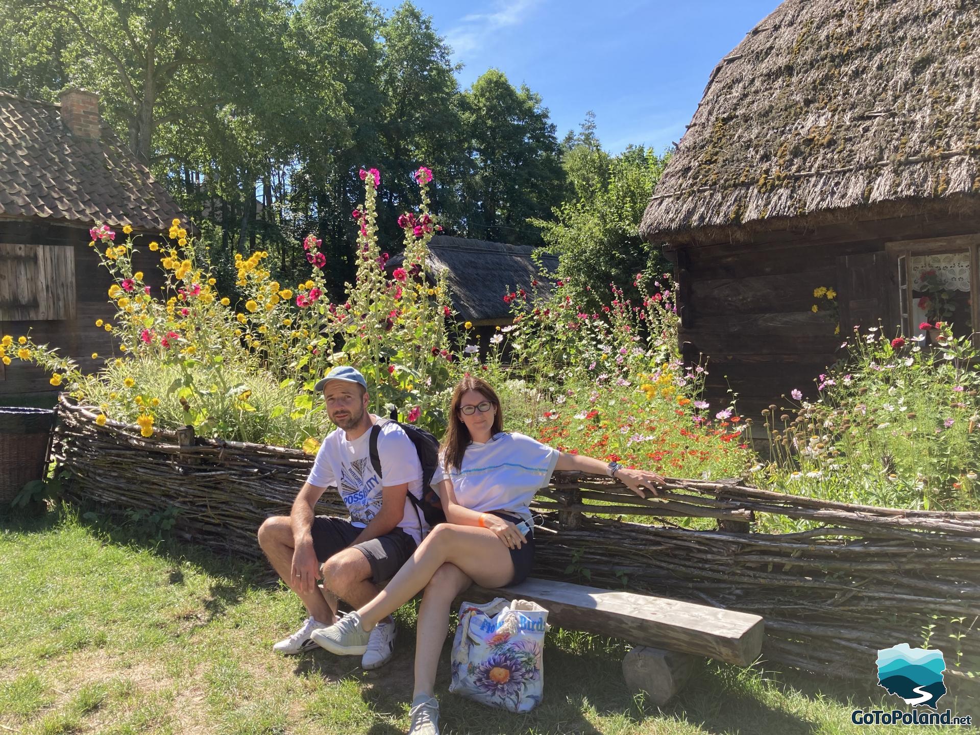 a man and a woman are sitting on a wooden bench, there are 3 huts behind them, wildflowers of different colors grow outside the huts