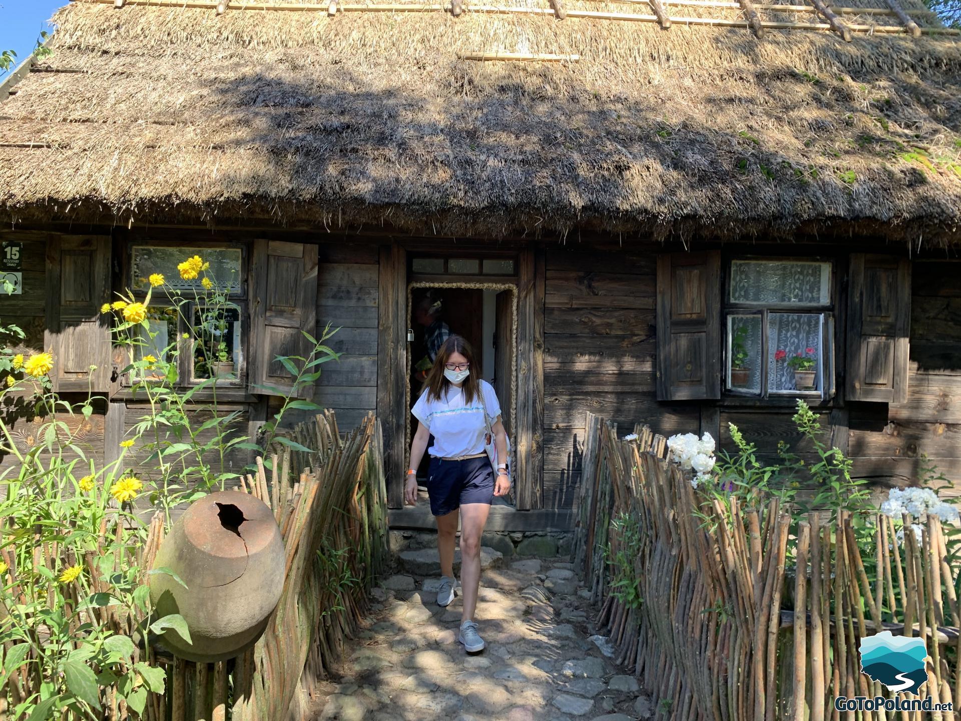 a woman is coming out of a peasant hut with thatched roof, on both sides of the path to the hut there is a fence made of branches