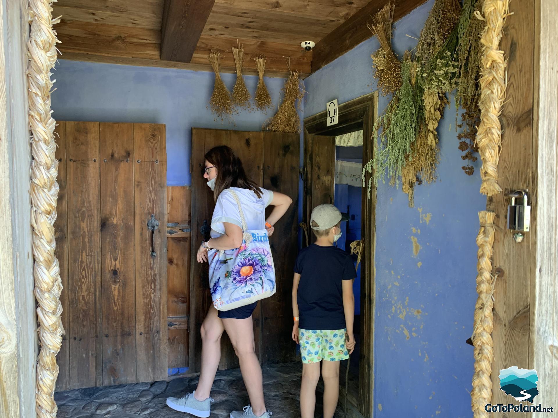 a woman and a child are in a peasants hut, dried herbs hanging from the low ceiling
