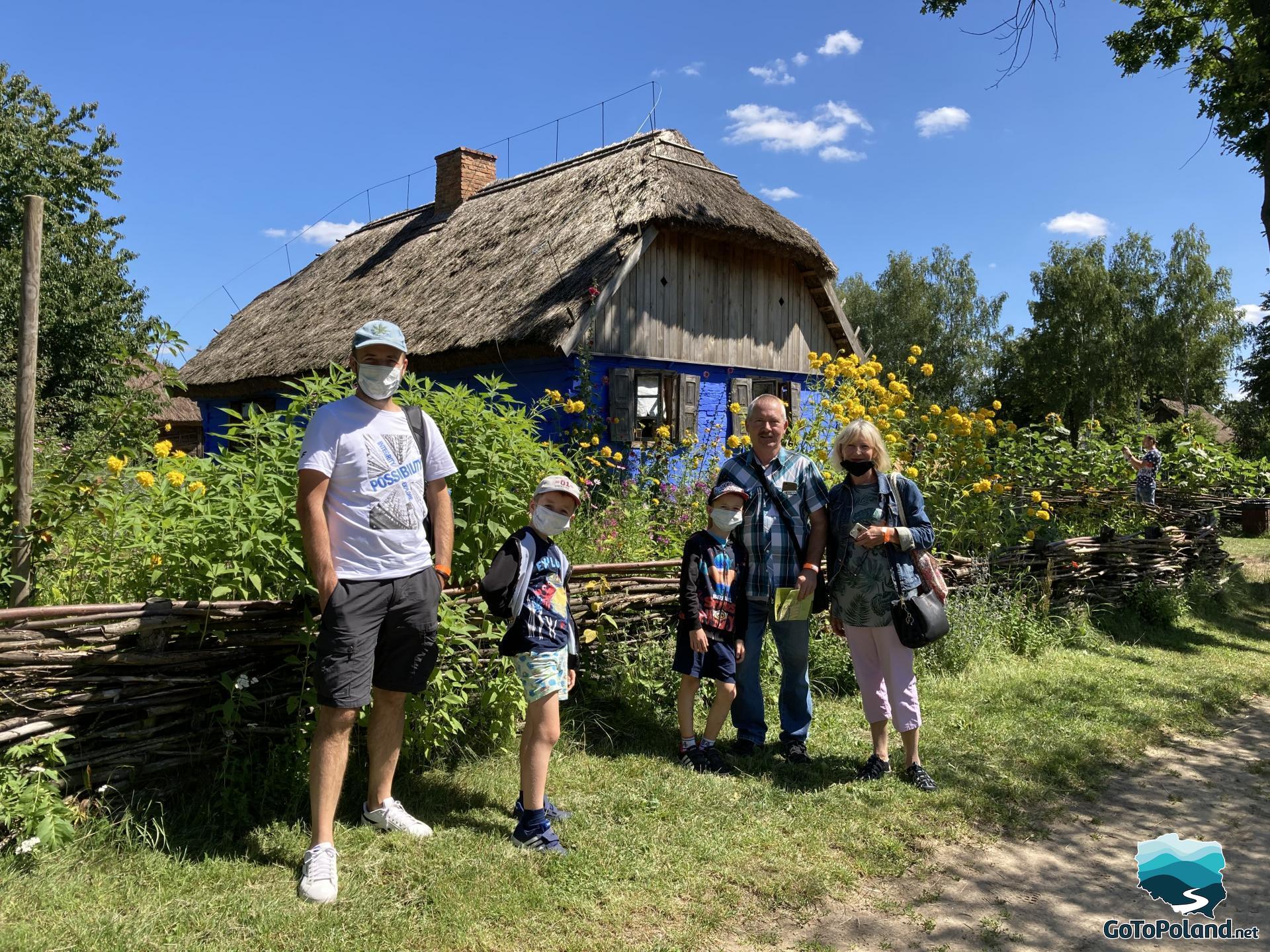 a family is standing in front of the blue hut, around the hut, there are lots of wild yellow flowers