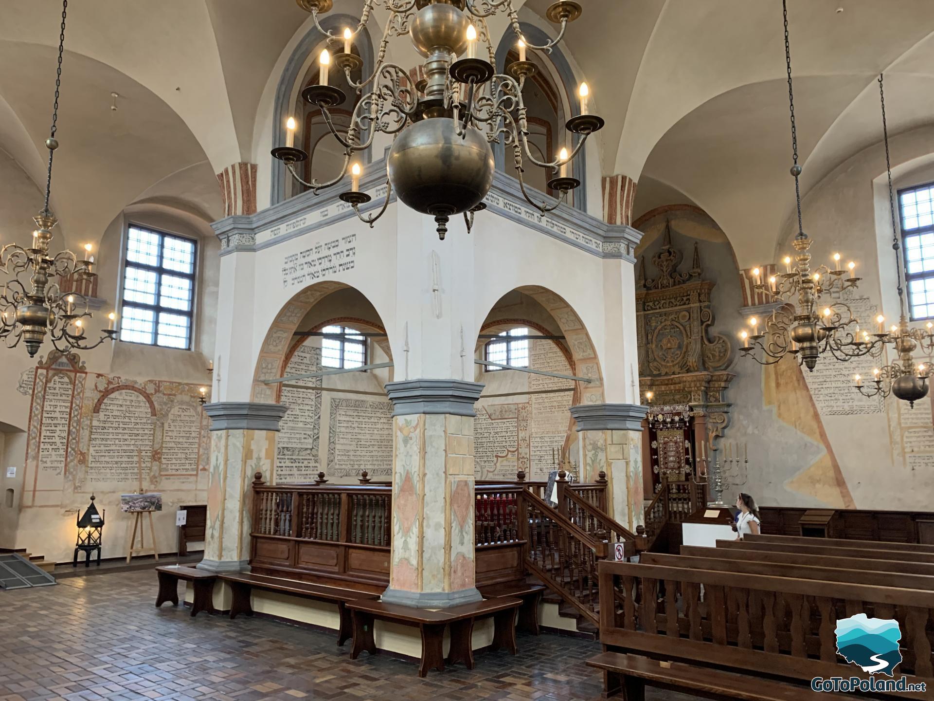 bimah in the synagogue, 4 chandeliers are lit, wooden benches, walls with Jewish inscriptions
