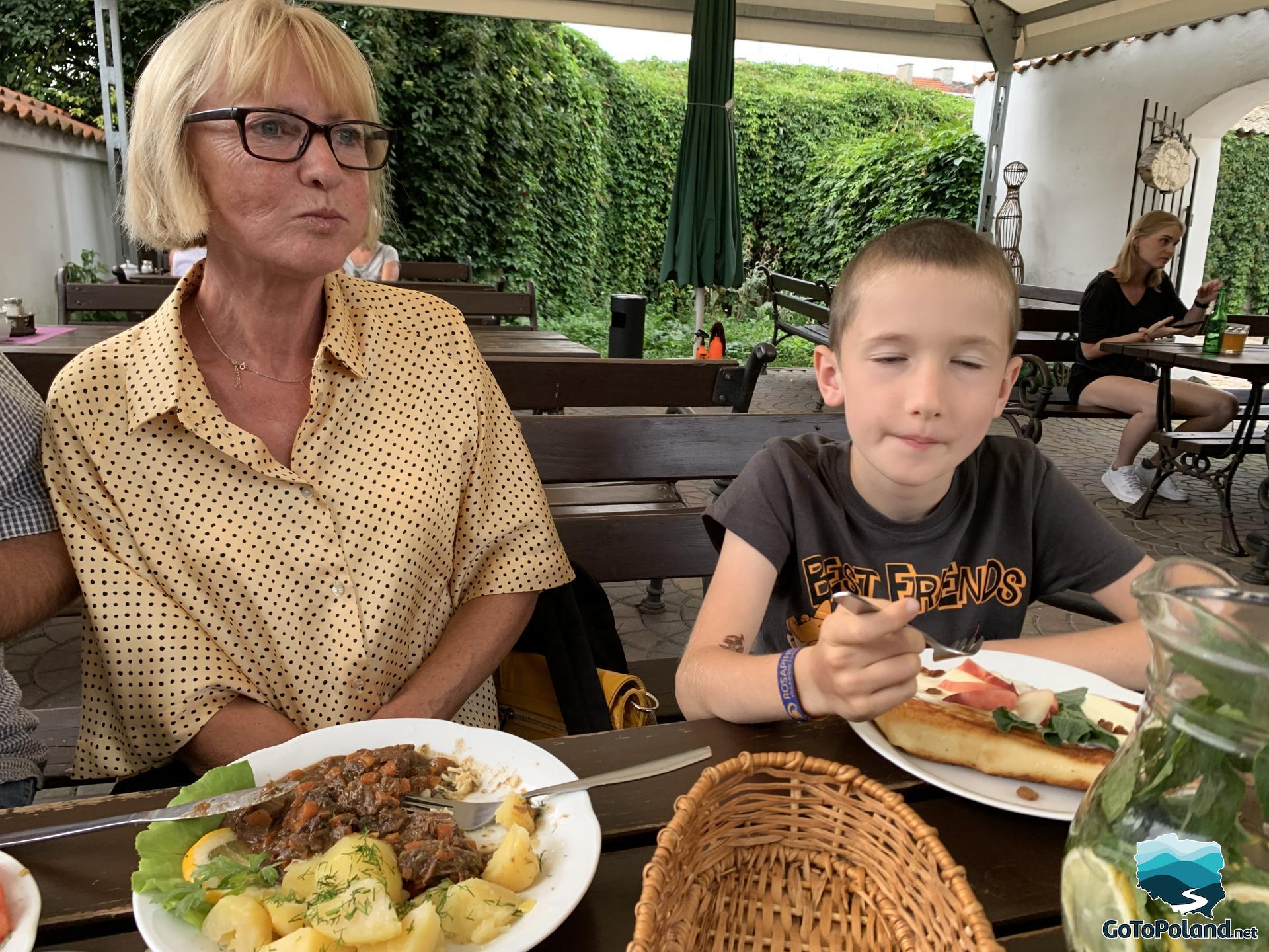 a woman and a boy are sitting at a table in a restaurant outdoors, the woman has potatoes and stew on a plate, the boy is eating pancakes, there is a jug with water, mint and lemons on the table