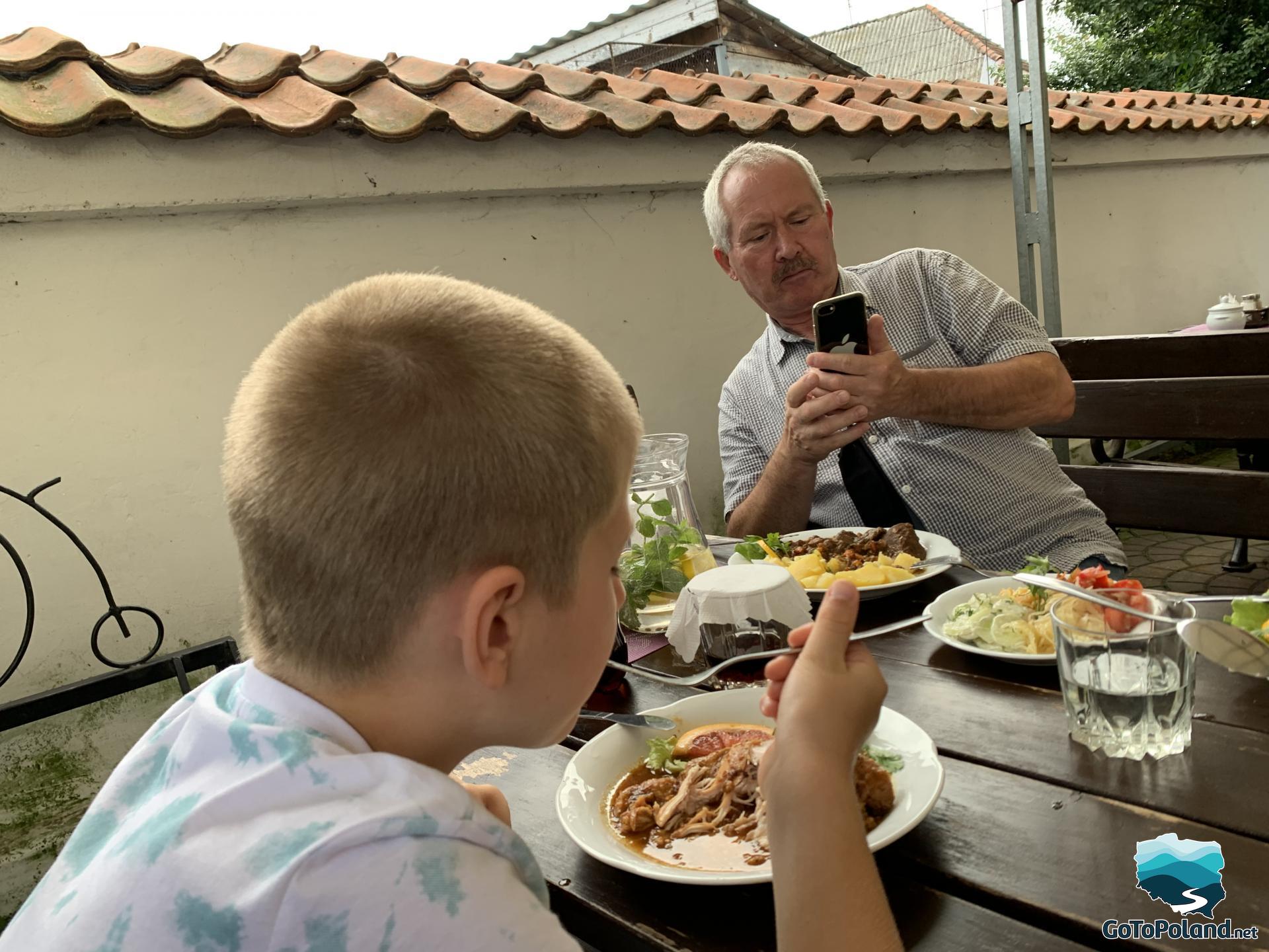 a boy is eating chicken in an outdoor restaurant, a man is taking a photo of the other dishes served on white plates