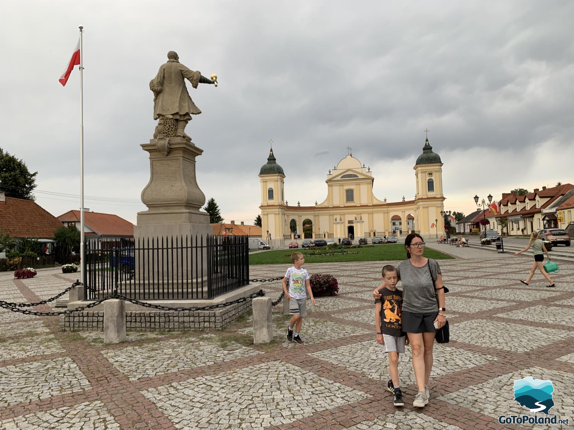 the main square in the town, a monument to a military commander, a woman and two children near the monument, a church with a yellow facade in the background
