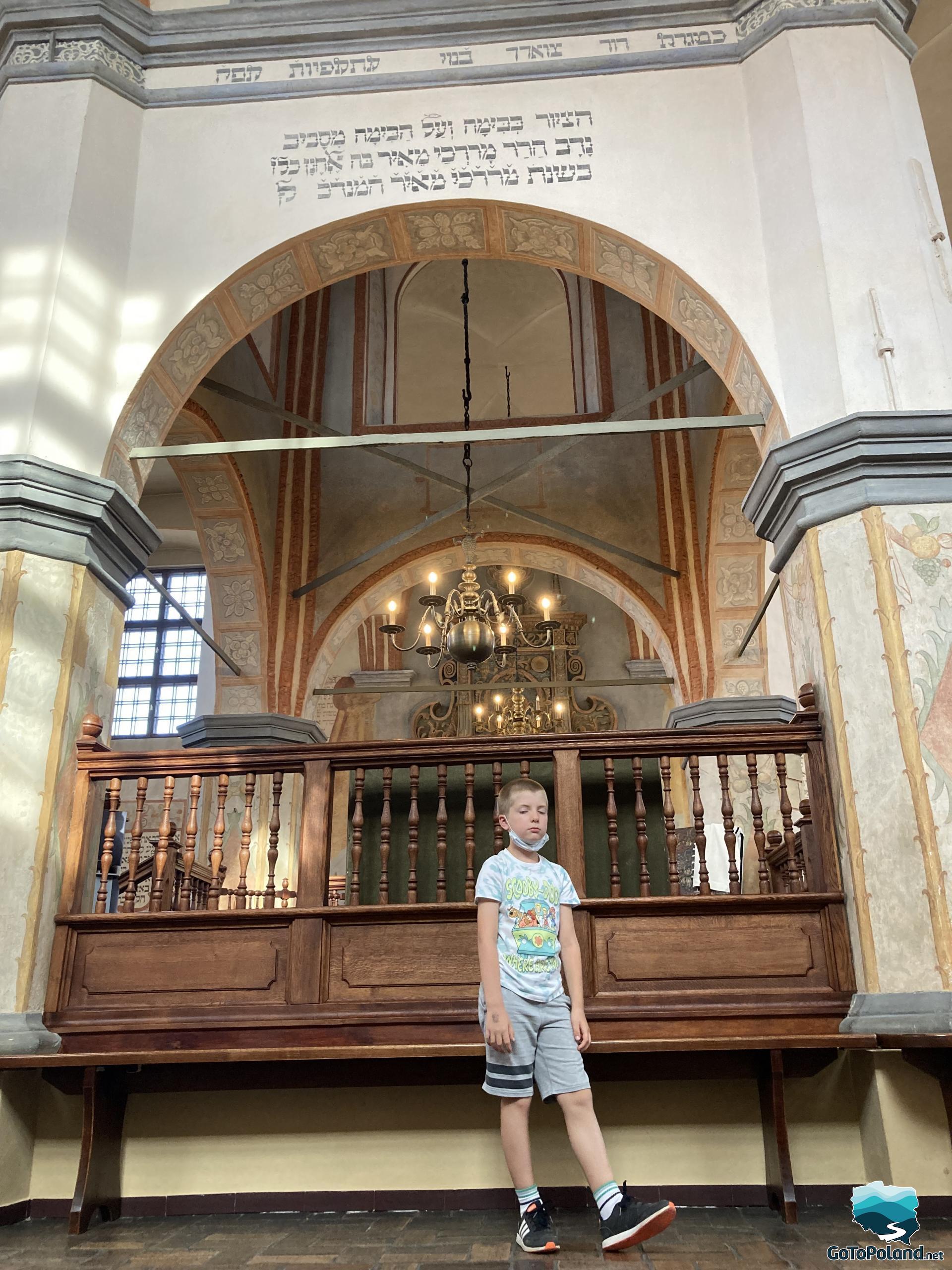 the boy is in the synagogue, he is standing by a wooden bench, there is an arch in the wall behind him, and an inscription in Yiddish above the arch