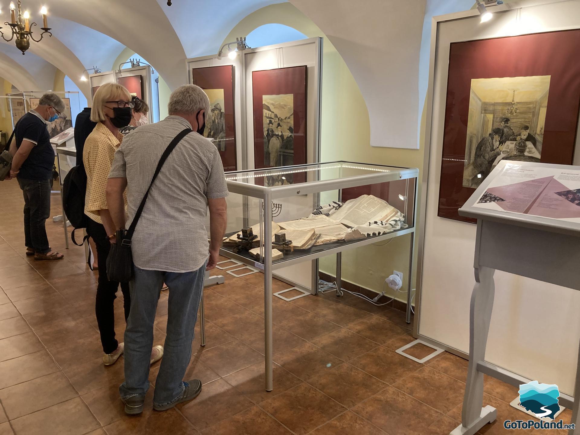 people are looking at the exhibits of Jewish culture that are in display cases, on the walls there are paintings depicting Jews in various activities