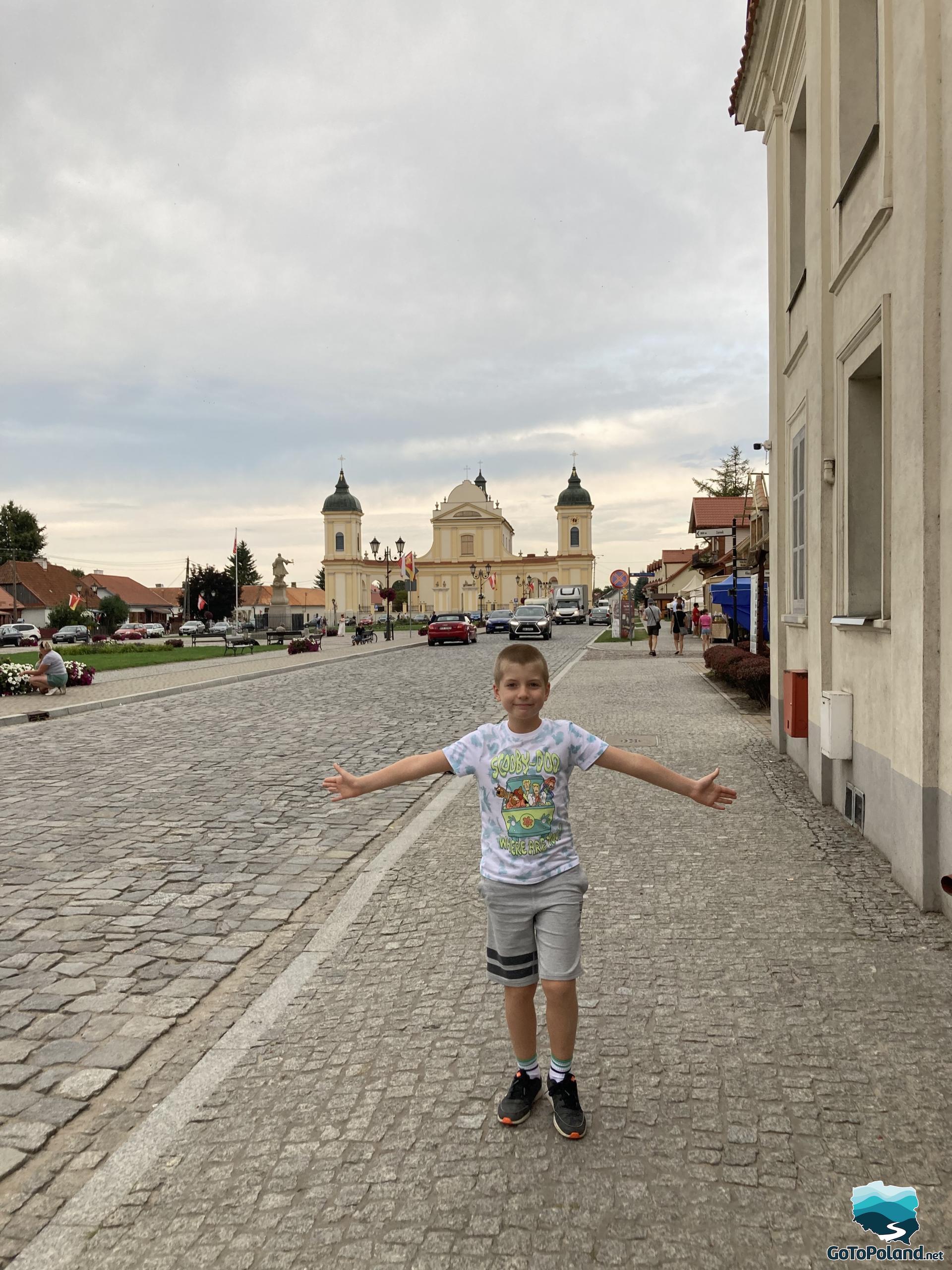 the boy is standing on the pavement, behind him the market square in the town and the yellow church in the background