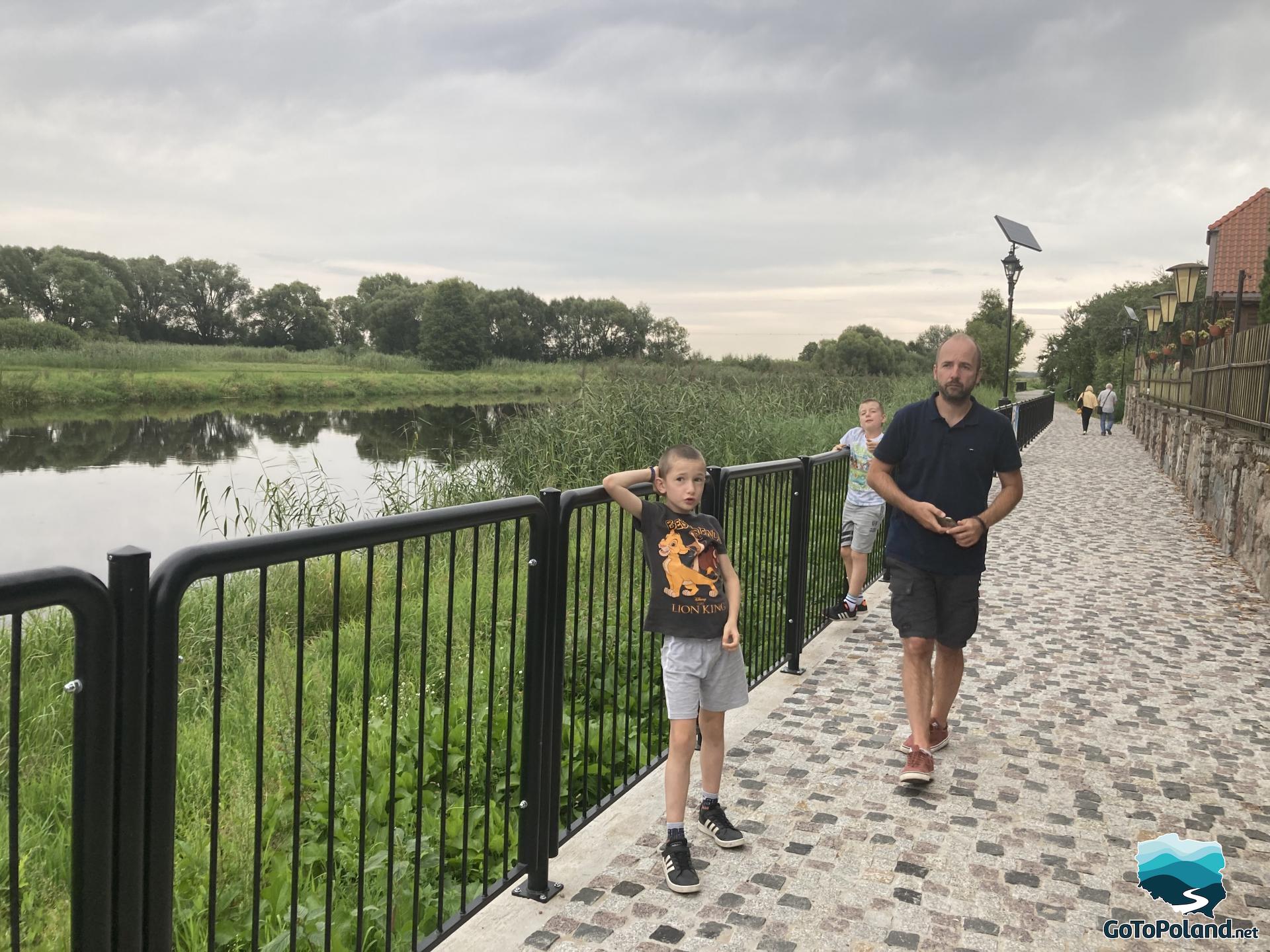 a man and two children on the pavement, a fence separates the pavement from the river, grass and reeds grow around the river