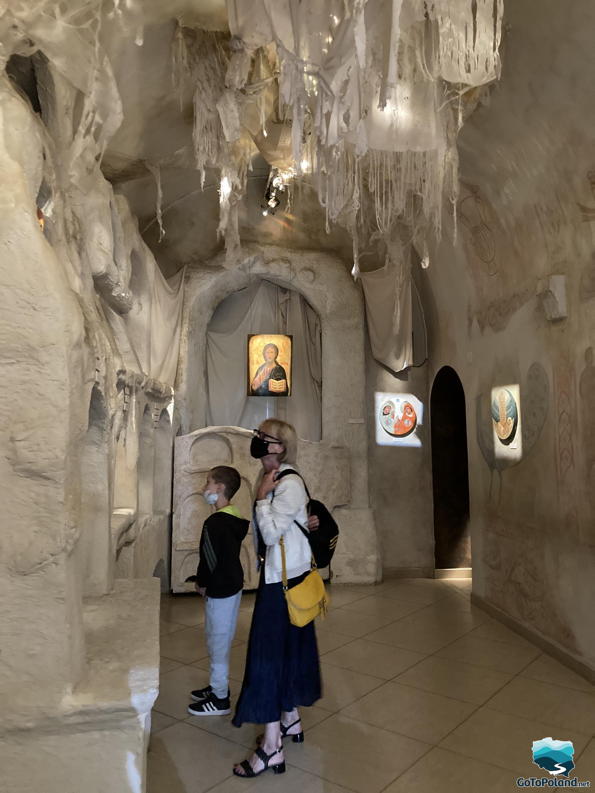 a woman and a boy are watching an exhibition of icons in a room stylized as a hermits cave